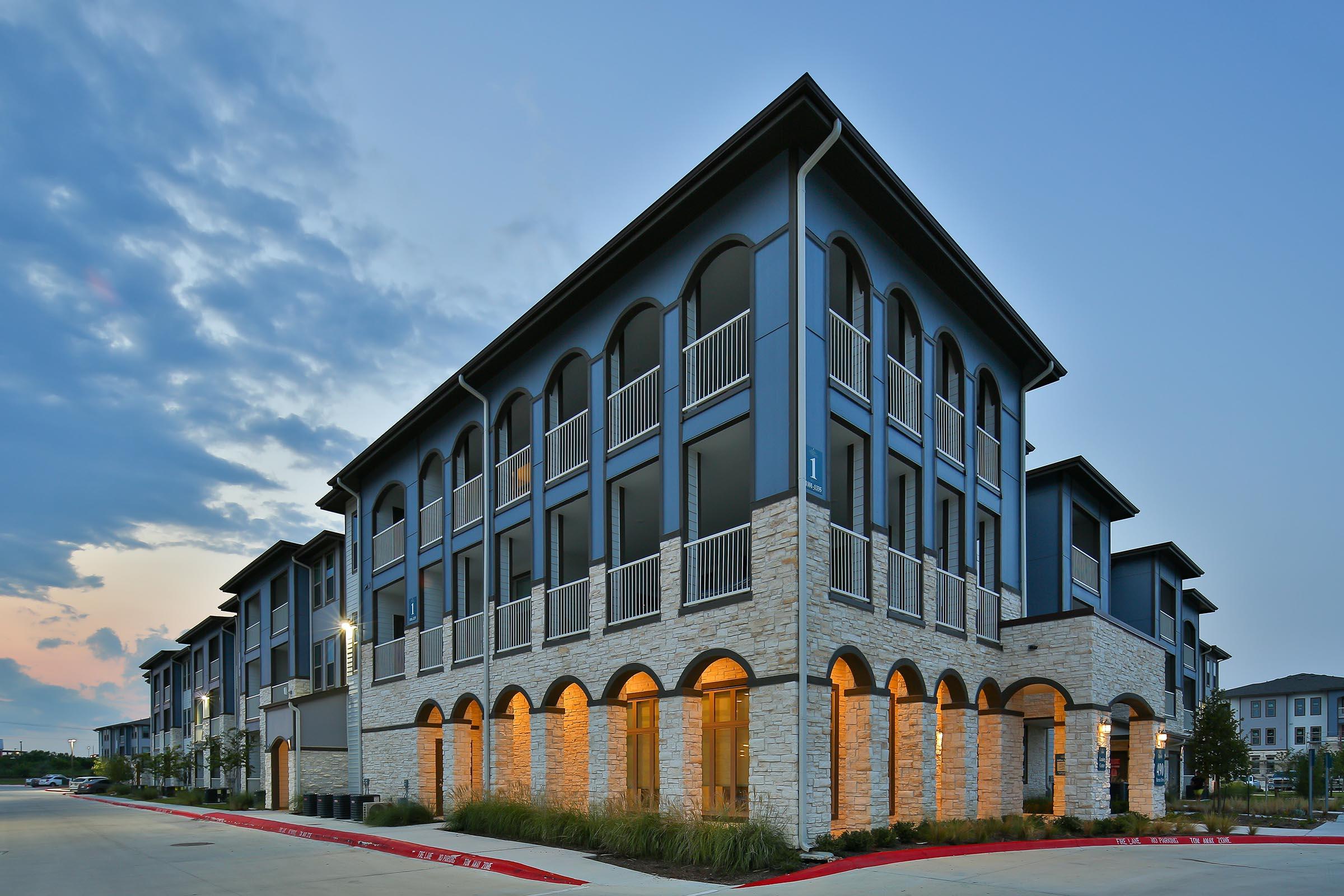 A modern multi-story apartment building with a combination of dark blue and stone exteriors. The structure features arched balconies and windows, with warm lighting illuminating the entryway. The scene is set during twilight, with a partly cloudy sky in the background.