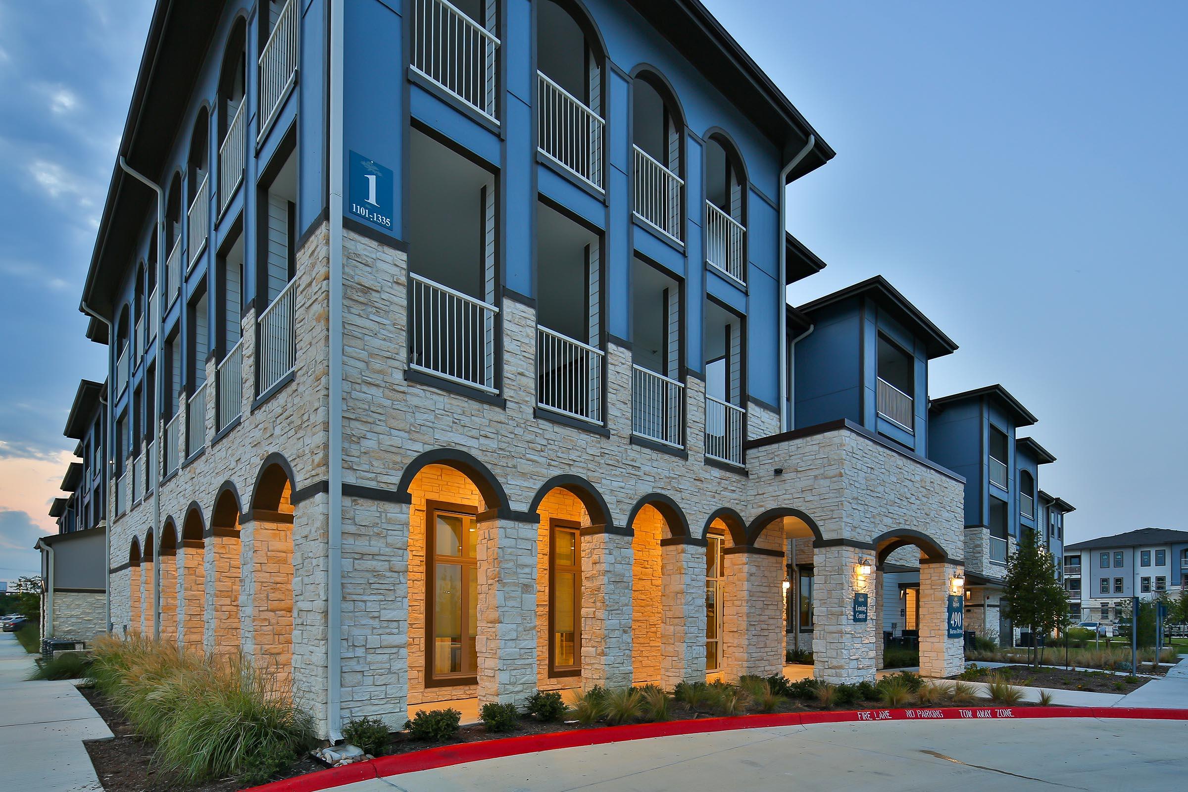 Modern multi-story residential building with a stone and blue exterior. The facade features arched entryways and balconies, illuminated by warm light. Surrounding landscaping includes shrubs and green grass, with a red curb marking the parking area. The sky above is partially cloudy, indicating either evening or early morning.
