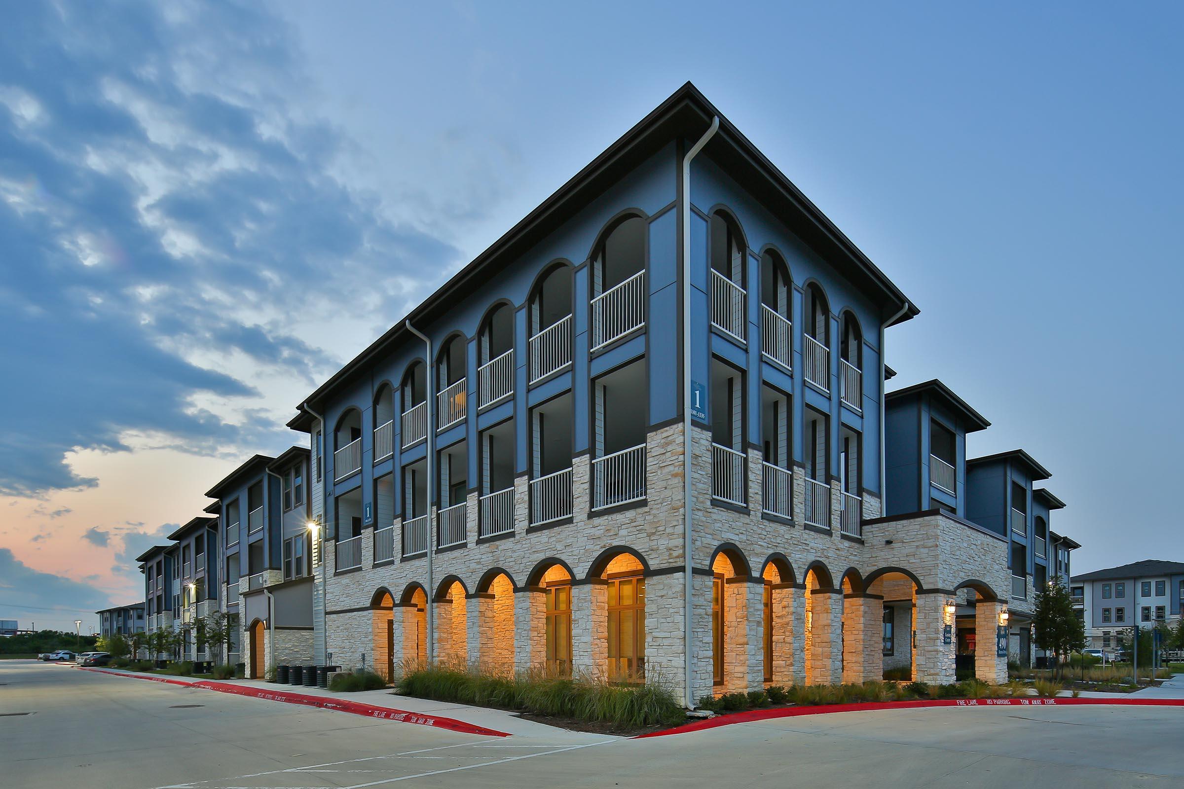 Modern multi-story apartment building with a blend of stone and contemporary design. The exterior features arched entryways and balconies. Soft evening light enhances the architectural details against a twilight sky, surrounded by landscaped greens and parking areas.