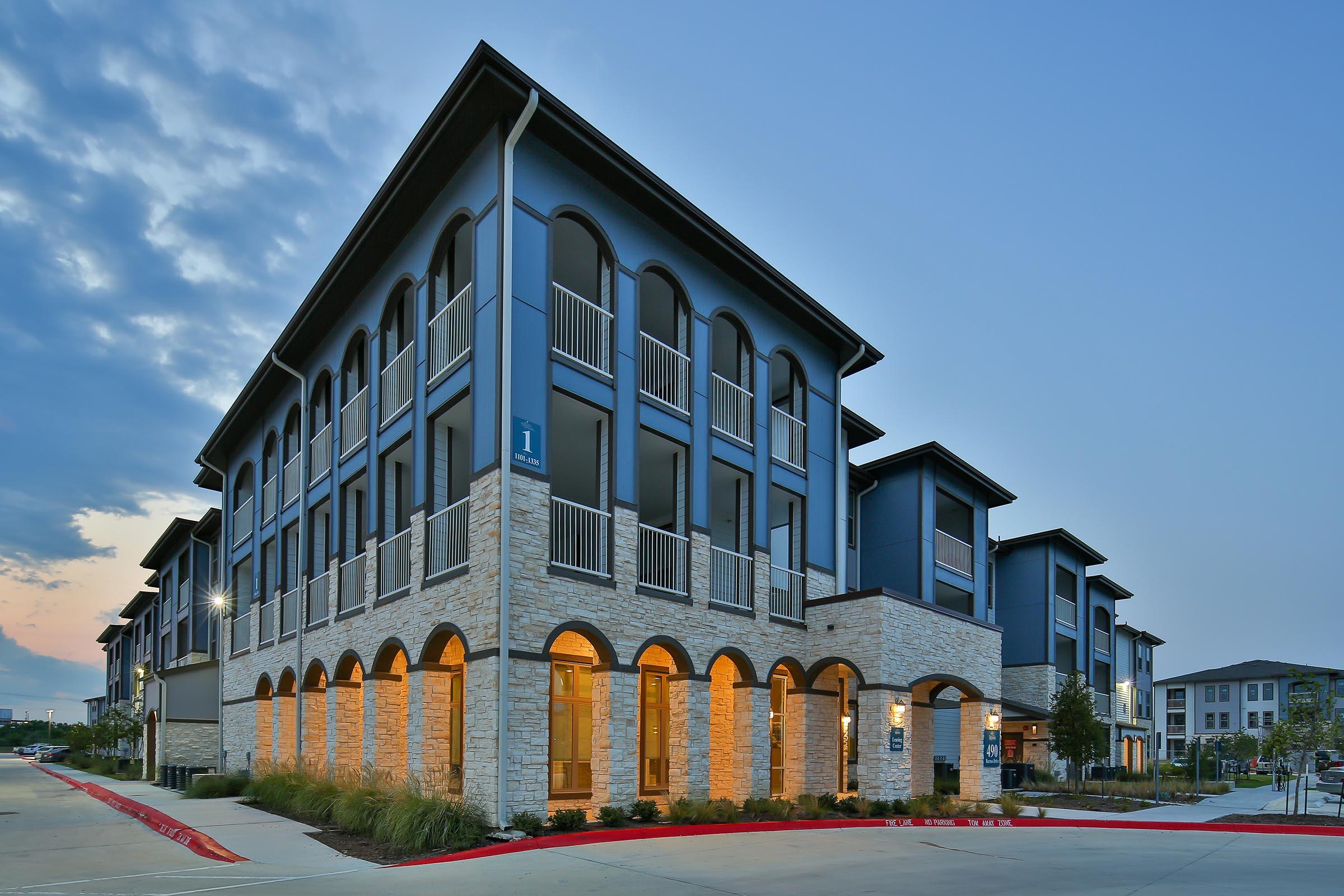 Modern apartment building with a contemporary design featuring stone and dark blue accents. The structure has arched entryways, multiple floors, and a well-lit facade. The setting is in the evening with soft lighting and a cloudy sky, showcasing the building's architectural details.