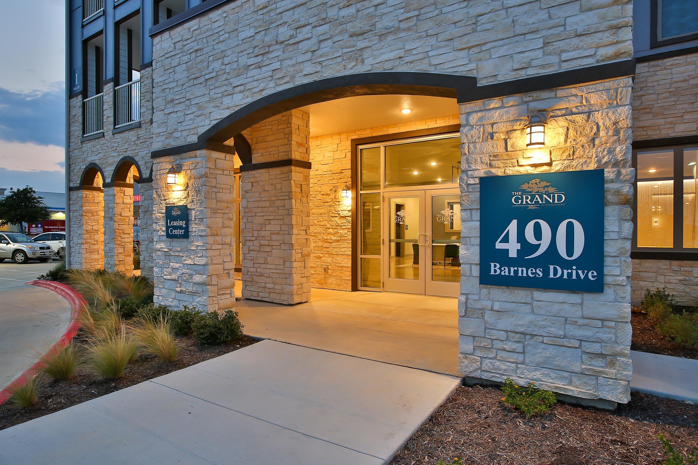 Entrance of a modern building featuring a stone facade and large glass doors. Signs display "The Grand" and the address "490 Barnes Drive." The surrounding area is landscaped with grass and plants, and the sky reflects a soft twilight glow.