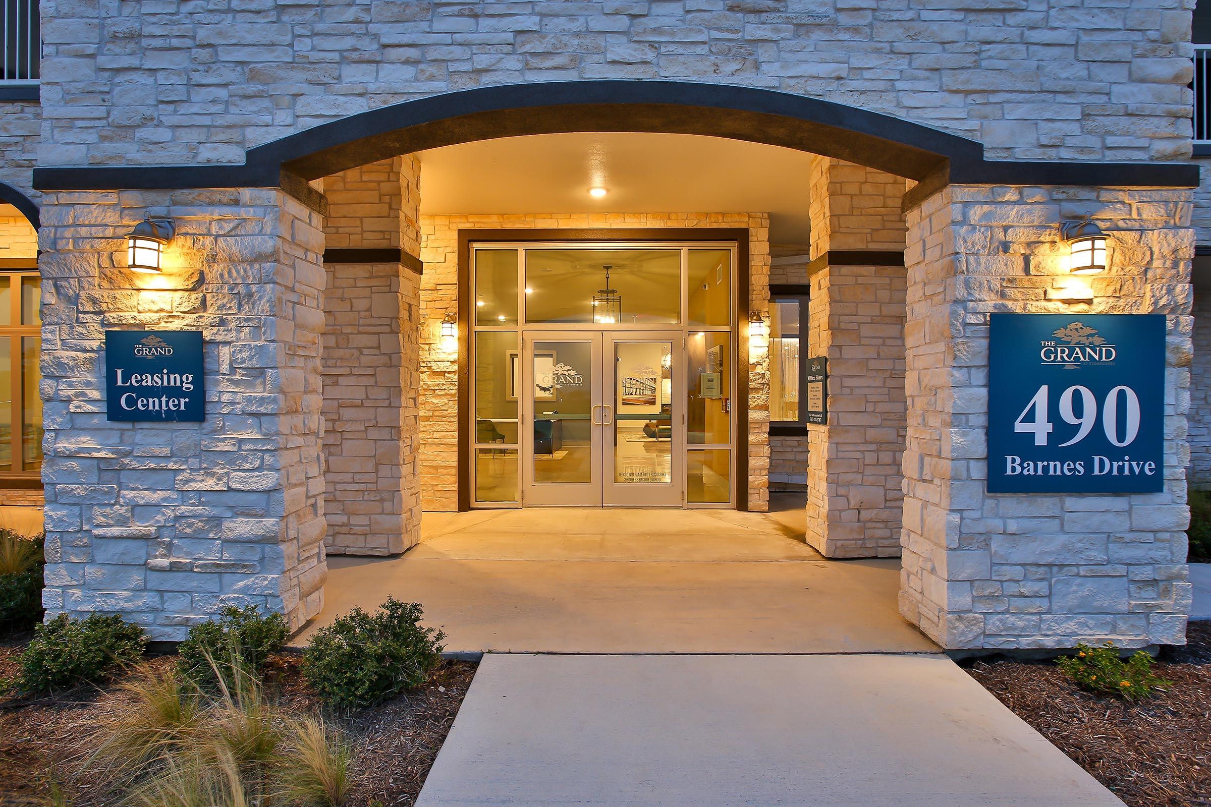 A stone facade of a leasing center entrance, featuring glass double doors, illuminated by wall sconces. Signs display "Leasing Center" and "490 Barnes Drive." Surrounding landscaping includes low shrubs, adding to the welcoming atmosphere.