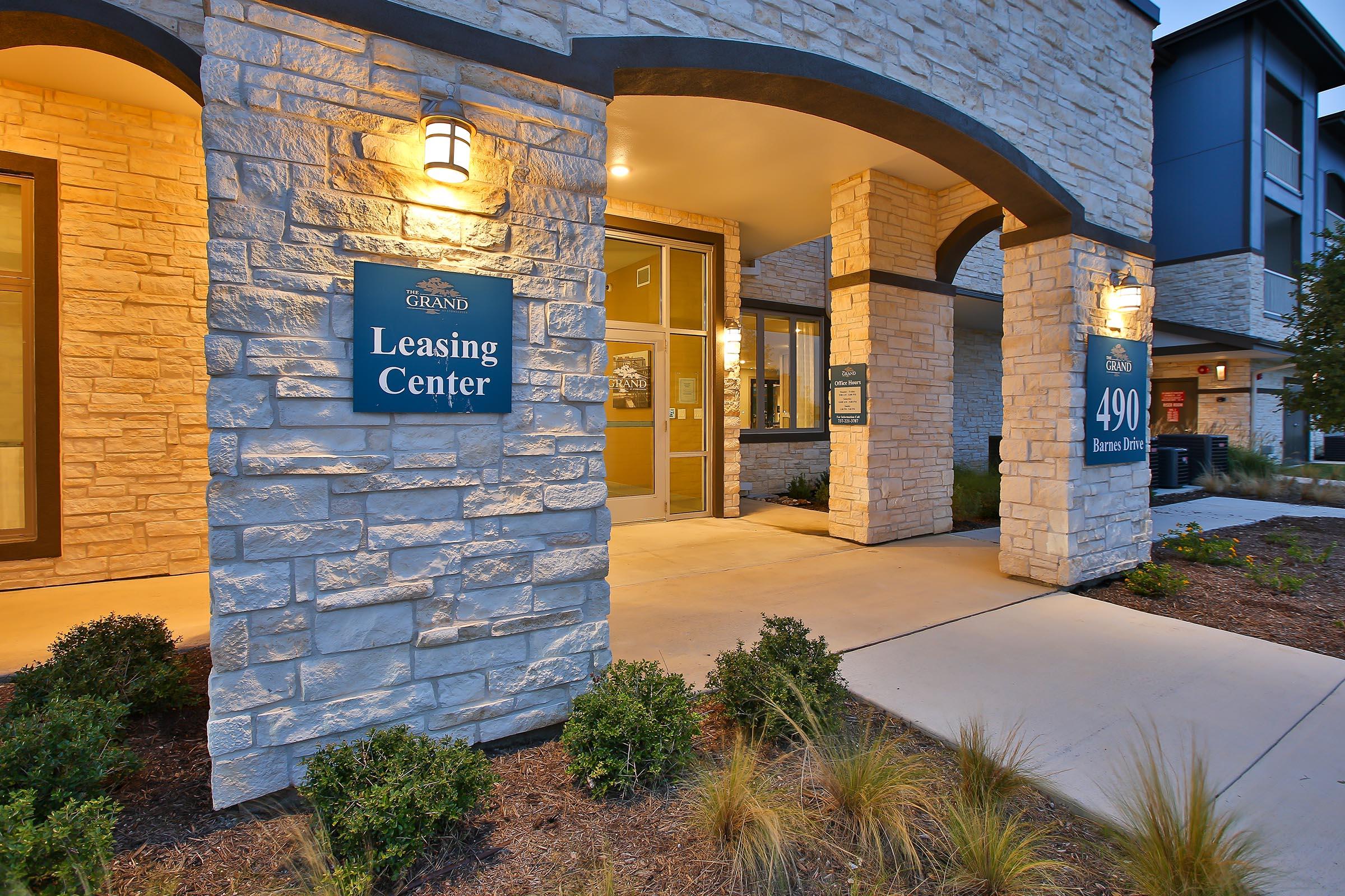 Exterior view of a leasing center with stone archways, illuminated signs featuring "Leasing Center" and the address "490 Barnes Drive." The entrance is framed by manicured landscaping, including shrubs and ornamental grasses, creating an inviting atmosphere.
