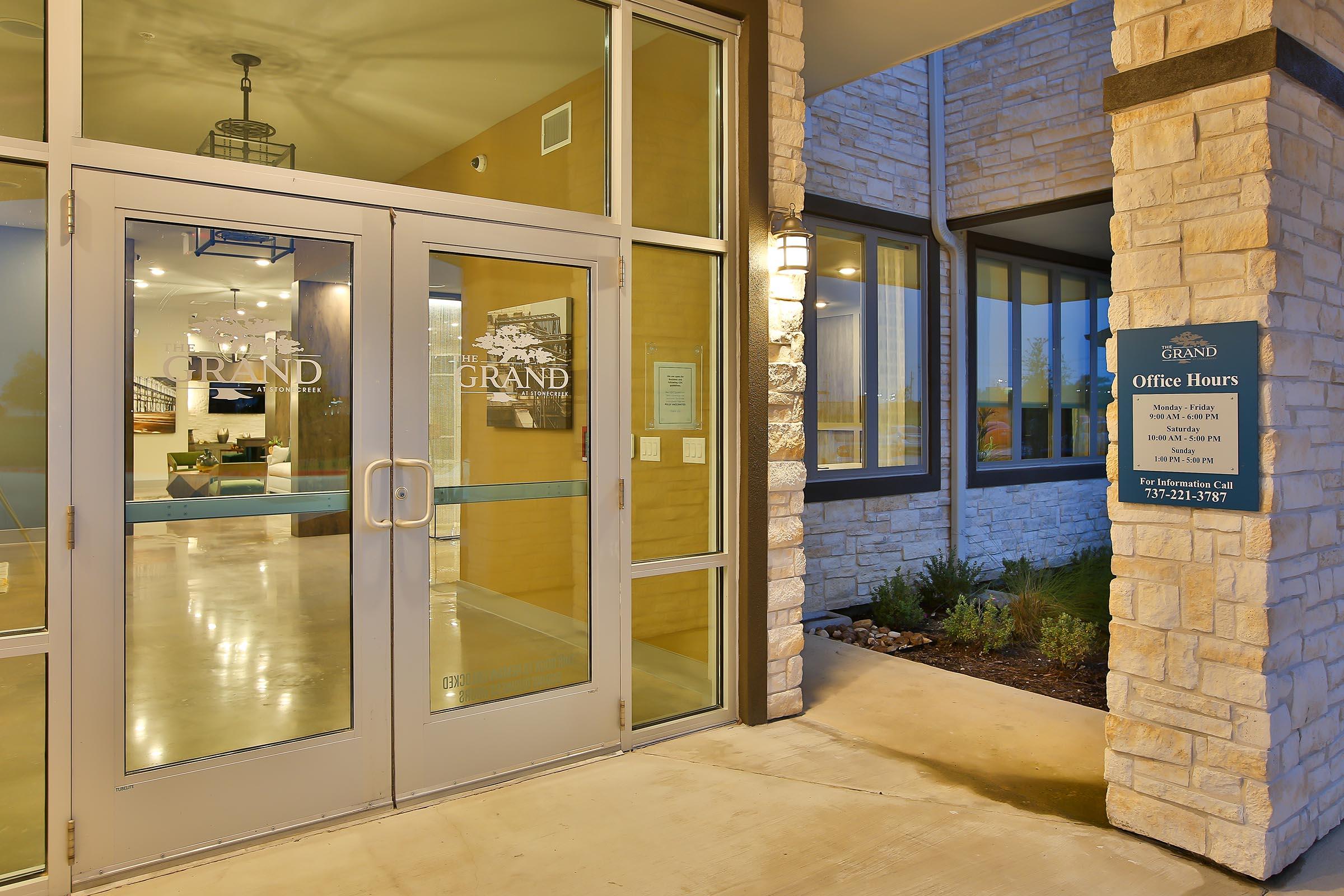Modern entrance of a building with large glass doors labeled "THE GRAND." Soft lighting and a stone-accented facade create an inviting atmosphere. A sign next to the entrance displays office hours and contact information, with well-maintained landscaping visible in the foreground.