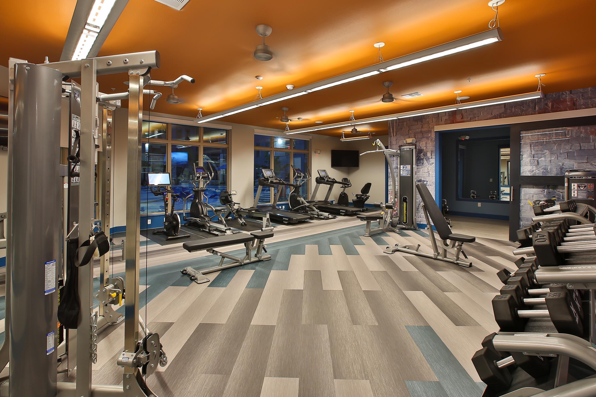 A modern gym interior featuring various exercise equipment, including weight machines, cardio machines, and free weights. The space has large windows allowing natural light, a ceiling with orange accents, and a clean, spacious layout with wooden flooring.