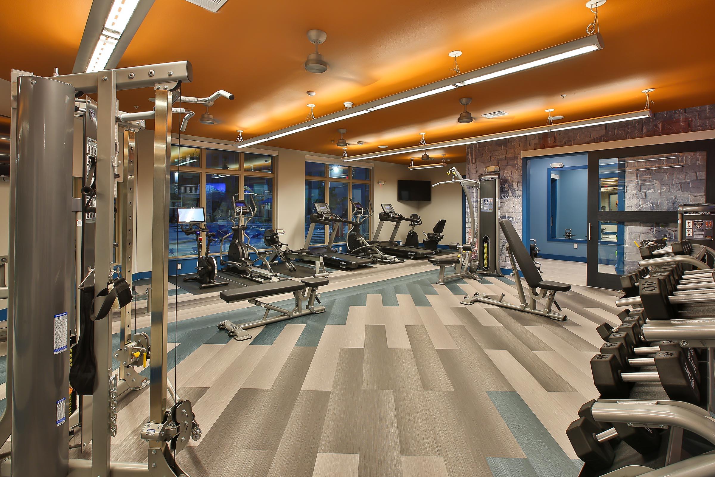 A modern gym interior featuring various exercise equipment, including weight machines and cardio machines. The space has large windows allowing natural light, with a stylish orange and gray color scheme, and is well-organized for a workout routine.