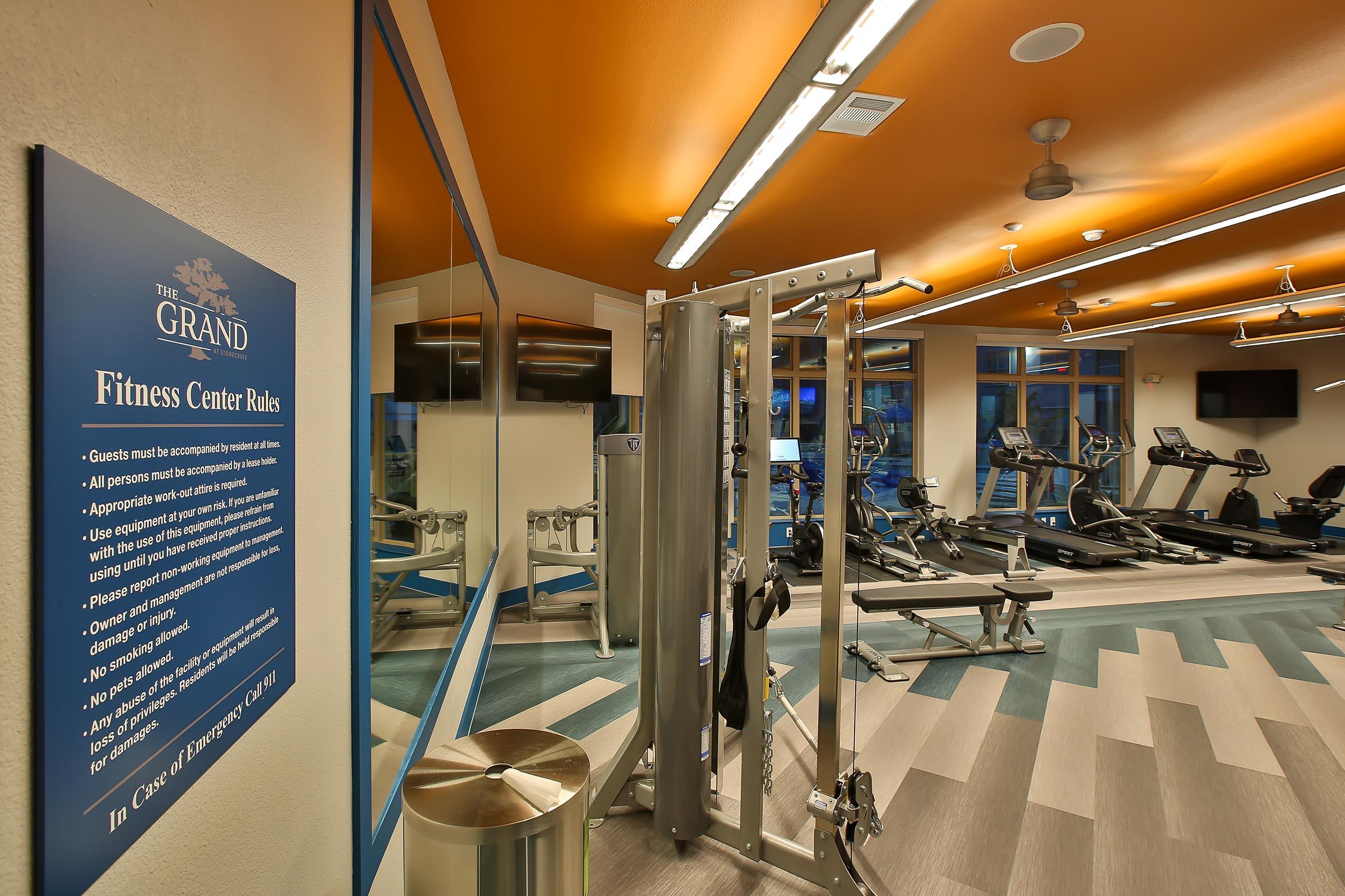 A modern fitness center featuring exercise equipment like treadmills and a weight machine. The wall displays fitness center rules. Brightly lit with an orange ceiling, large mirrors, and a clean, open layout, it emphasizes a welcoming atmosphere for guests.