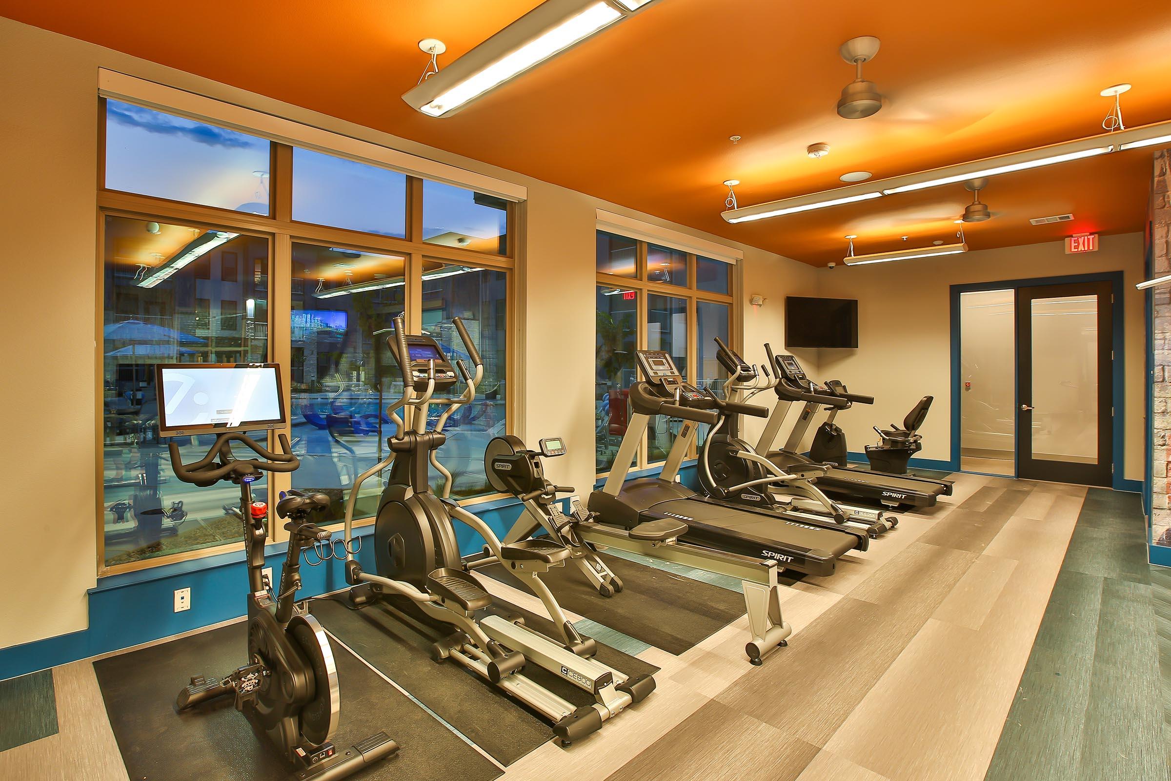 A modern gym interior featuring various exercise equipment including treadmills, elliptical machines, and stationary bikes, with large windows allowing natural light and a vibrant orange ceiling. The space is clean and well-lit, creating an inviting atmosphere for fitness activities.