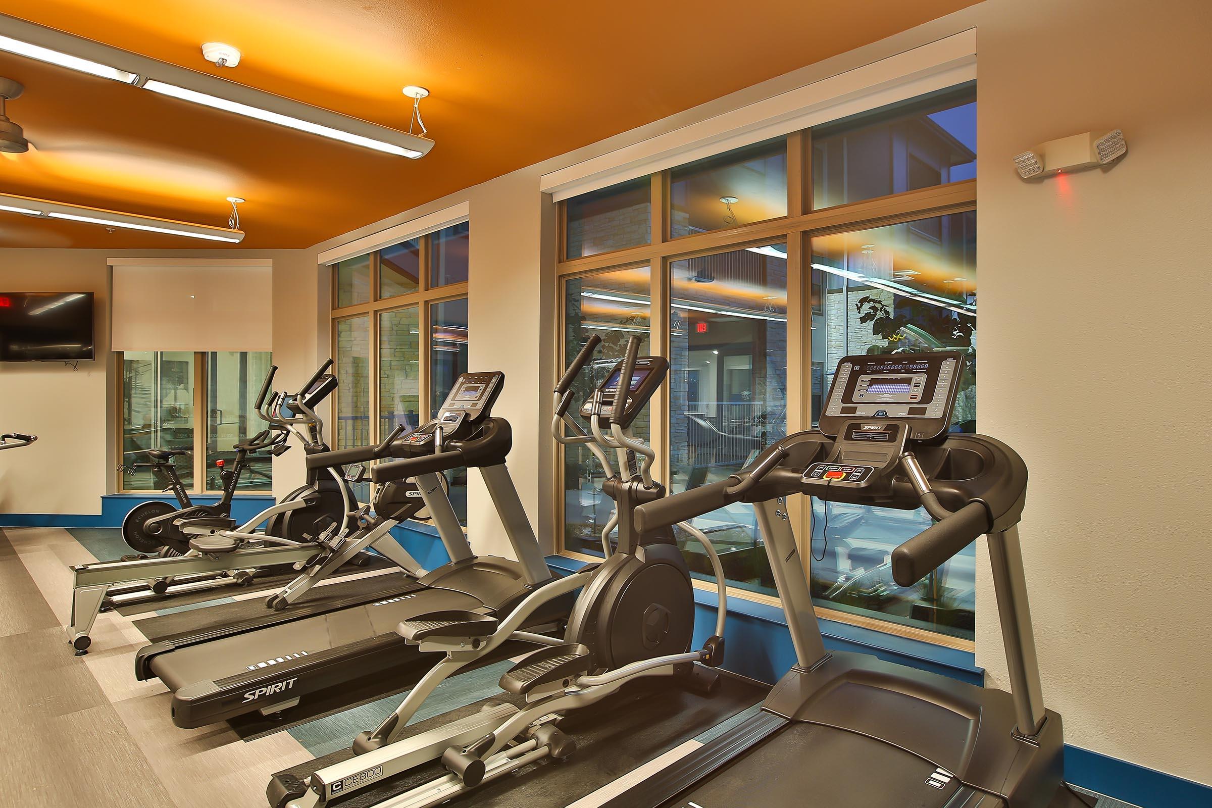 A modern gym interior featuring three exercise machines: two treadmills and one elliptical. Large windows let in natural light, illuminating the space with an orange and blue color scheme. Wall-mounted TV visible, creating an inviting workout environment.