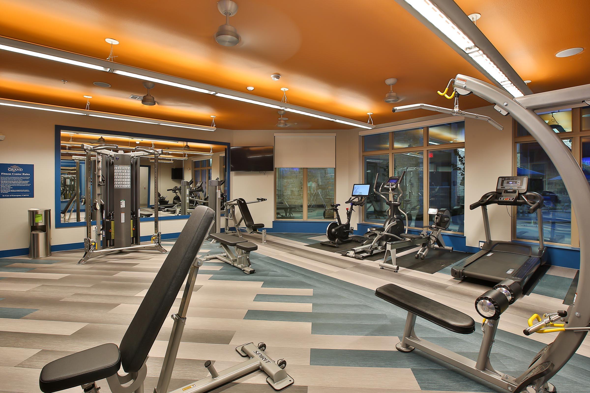 A modern gym interior featuring various exercise equipment, including weight machines, a treadmill, and an elliptical. The space is well-lit with large windows, blue and orange accents, and a clean, inviting atmosphere, designed for fitness enthusiasts.