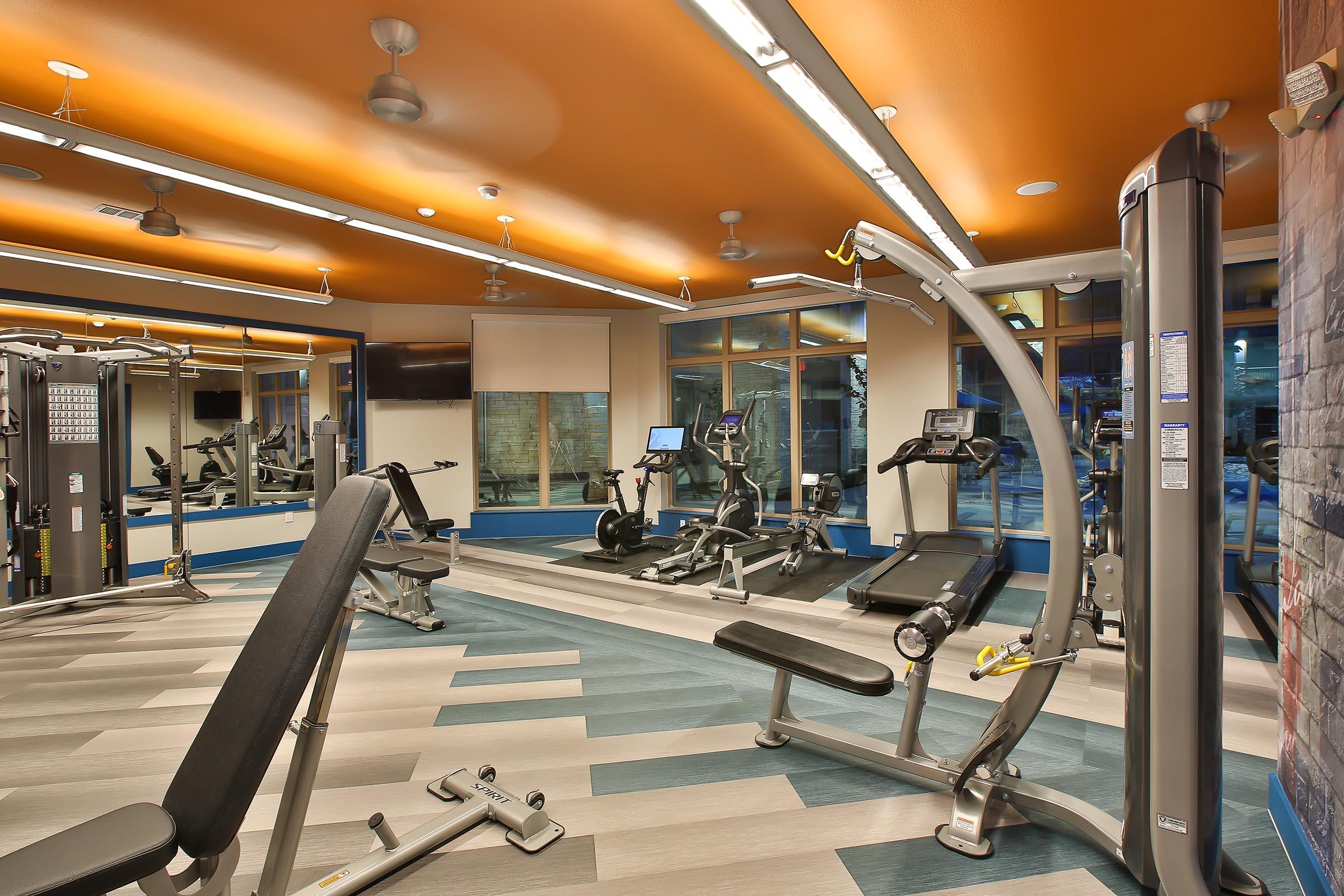 A modern gym interior featuring various exercise equipment, including weight machines and cardio machines, under bright lighting. The walls are painted in orange and blue, creating a vibrant atmosphere. Large windows provide natural light, enhancing the overall spacious feel of the room.