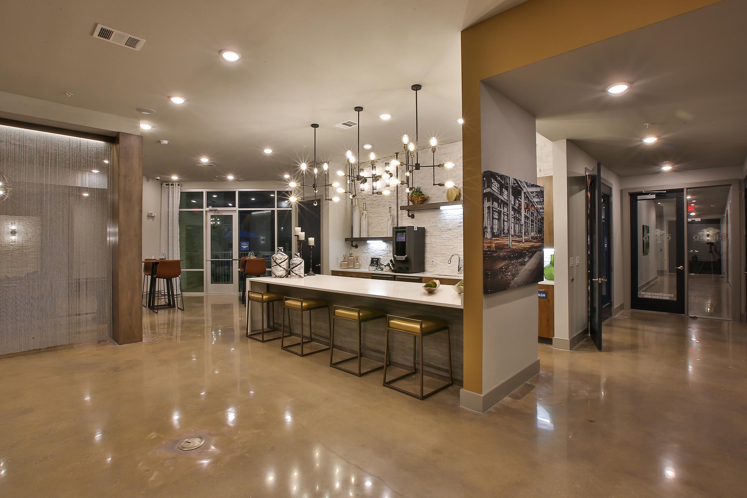 Modern kitchen and living area featuring a large island with gold bar stools, pendant lights, and stylish decor. An open layout showcases polished concrete flooring and contemporary furnishings, with large windows allowing natural light. A cozy dining nook is visible in the background, enhancing the inviting atmosphere.