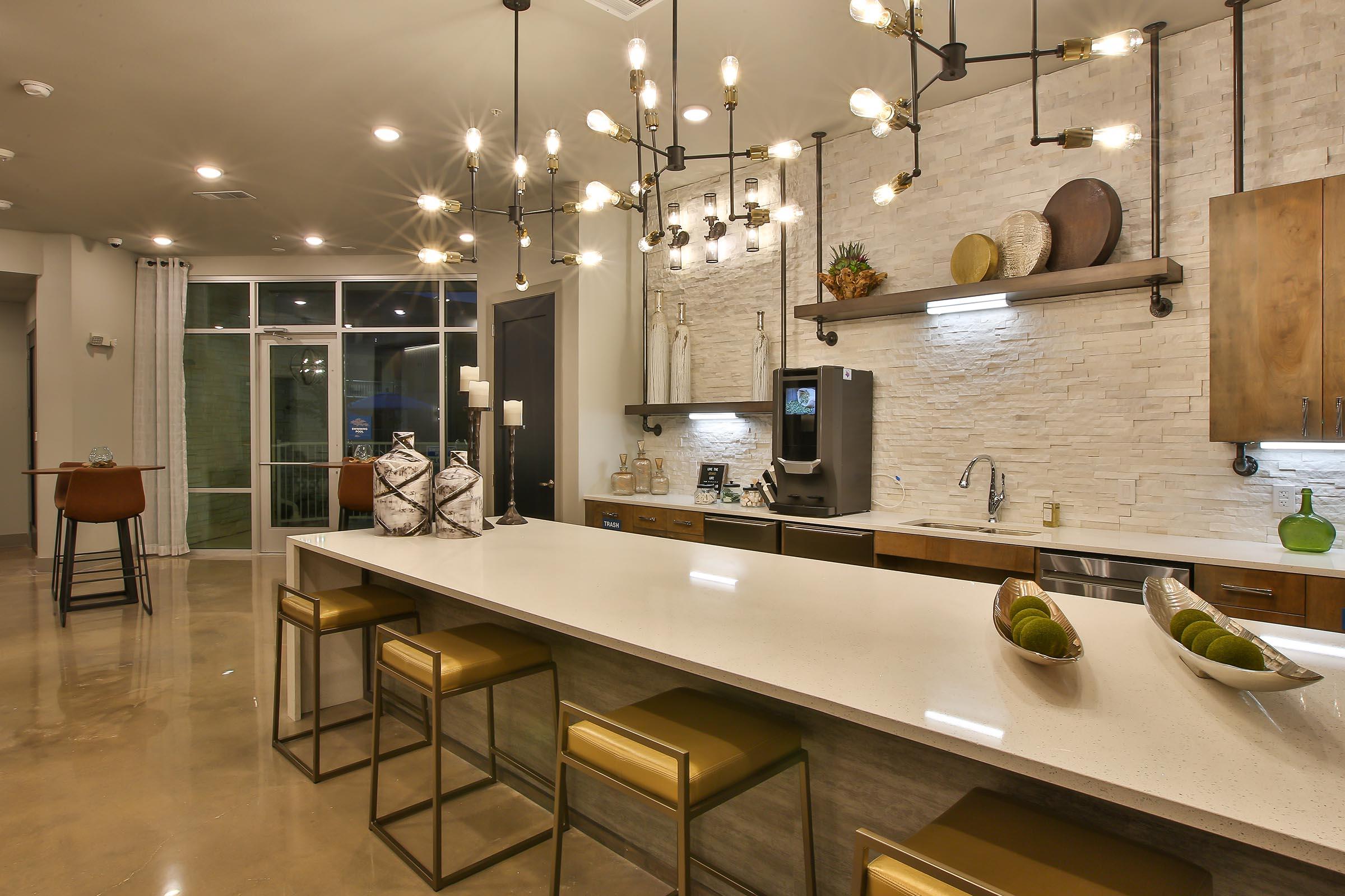 Modern kitchen with a large island bar featuring stylish stools, elegant lighting fixtures, and decorative accessories. The backsplash is made of light-colored stone, and there are kitchen appliances, including a coffee machine. A large window provides natural light, and there are greenery accents throughout the space.