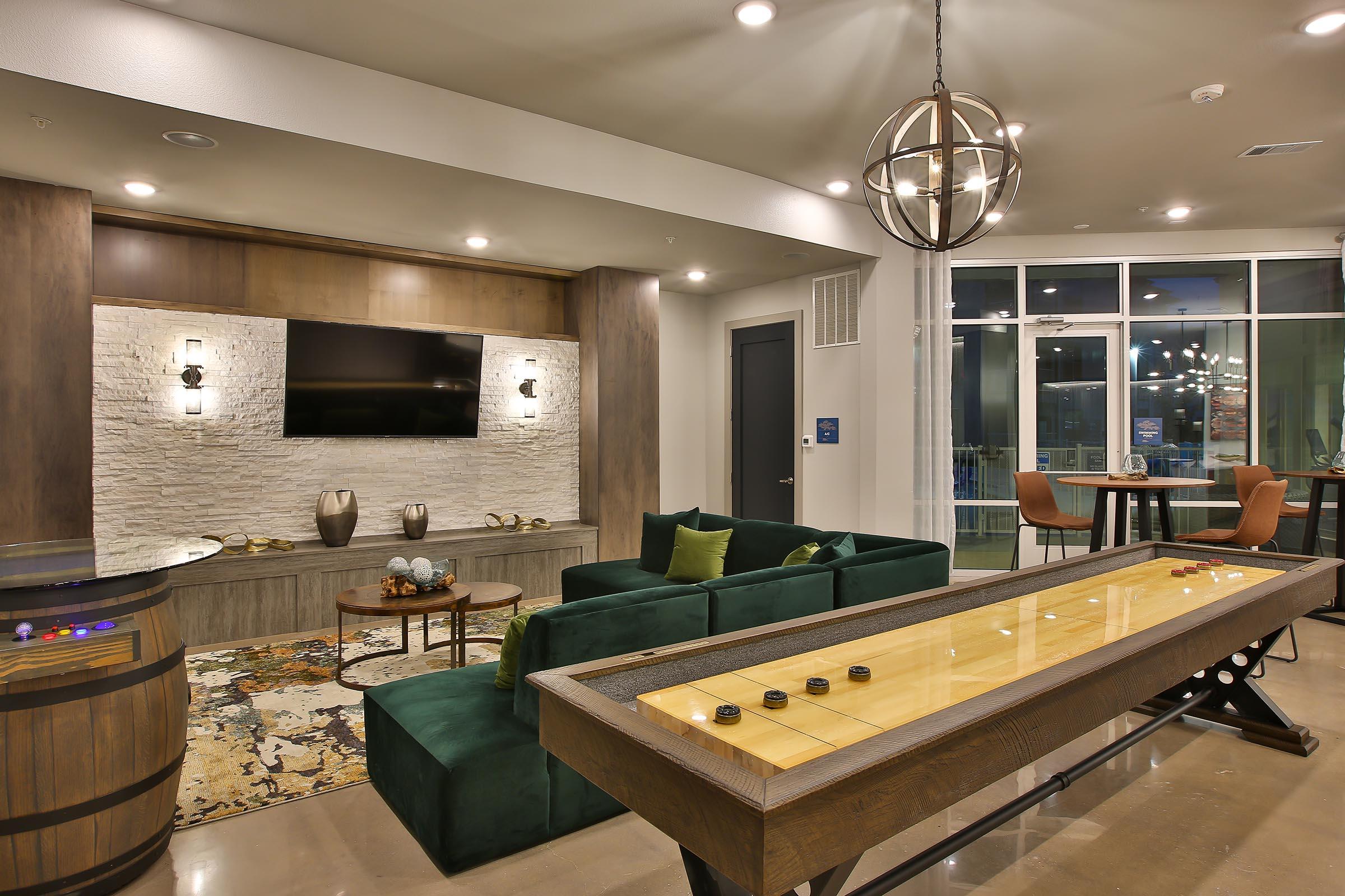 A modern lounge area featuring a green sectional sofa, a wooden shuffleboard table, and decorative vases on a stone wall. A large flat-screen TV is mounted above a stylish side table, with soft lighting and a cozy ambiance complemented by a dining area in the background.