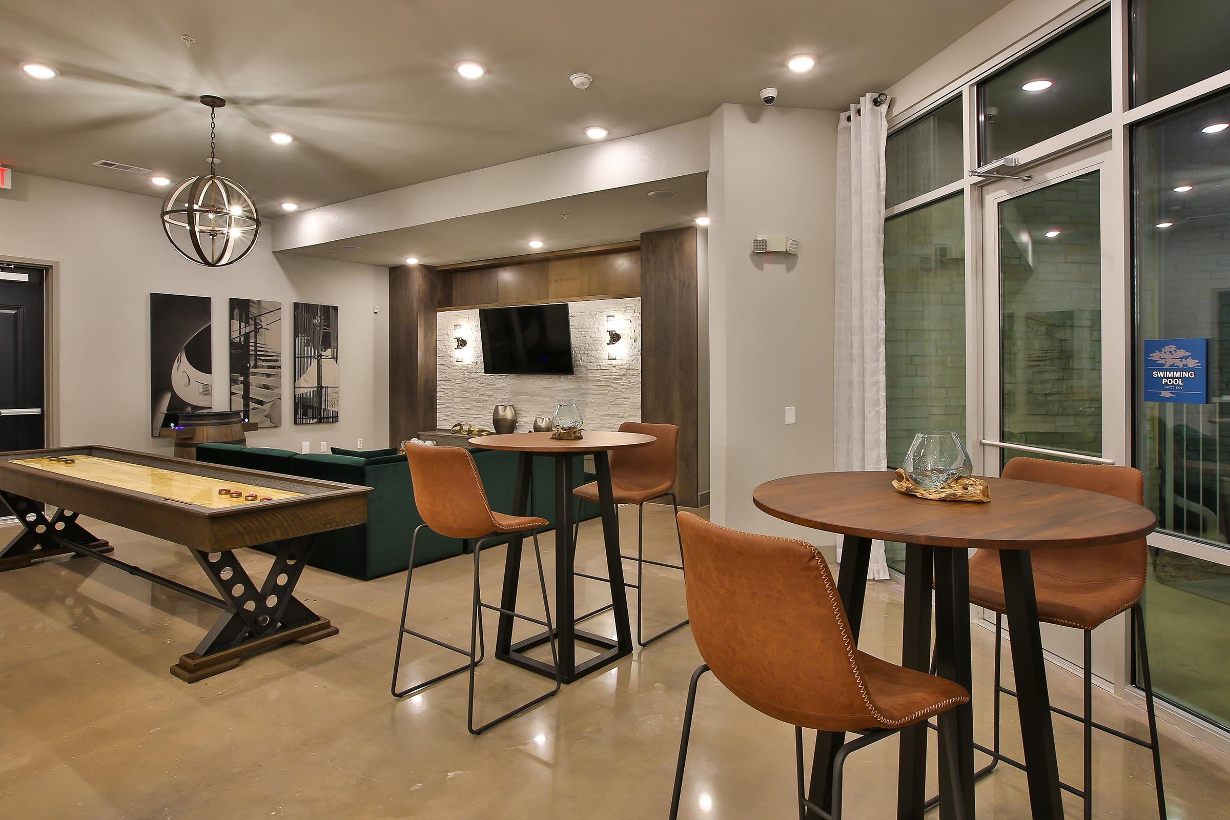 A modern lounge area featuring two tall tables with brown leather stools, a shuffleboard table, and a large TV mounted on a stone wall. The room has stylish lighting and large windows allowing natural light, creating a welcoming atmosphere for social gatherings.