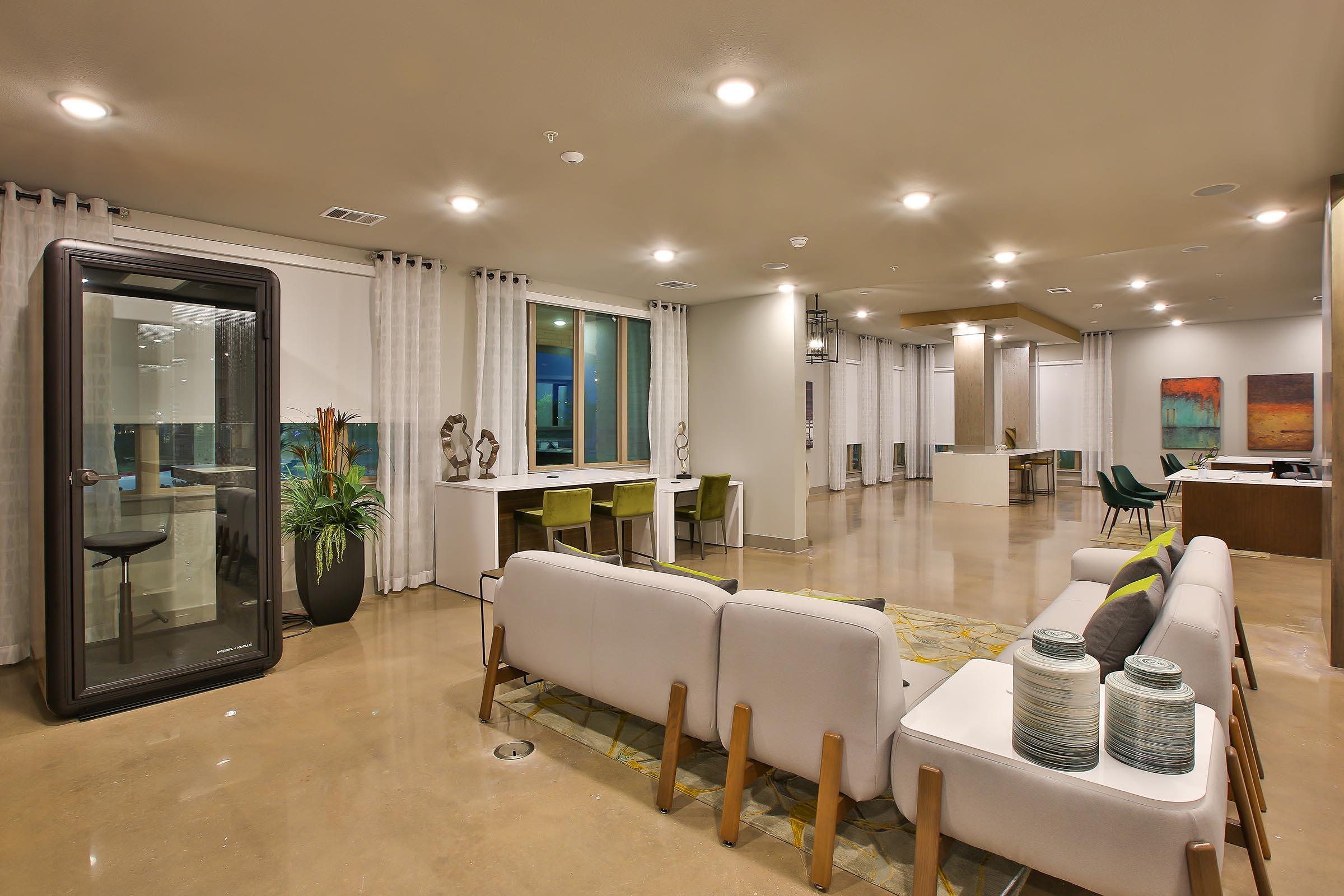 Modern living space featuring large windows with natural light, a comfortable seating area with a sofa, decorative vases on a table, a desk setup, and a glass-enclosed meeting pod. The room has light-colored walls, polished concrete floors, and contemporary artwork on display.