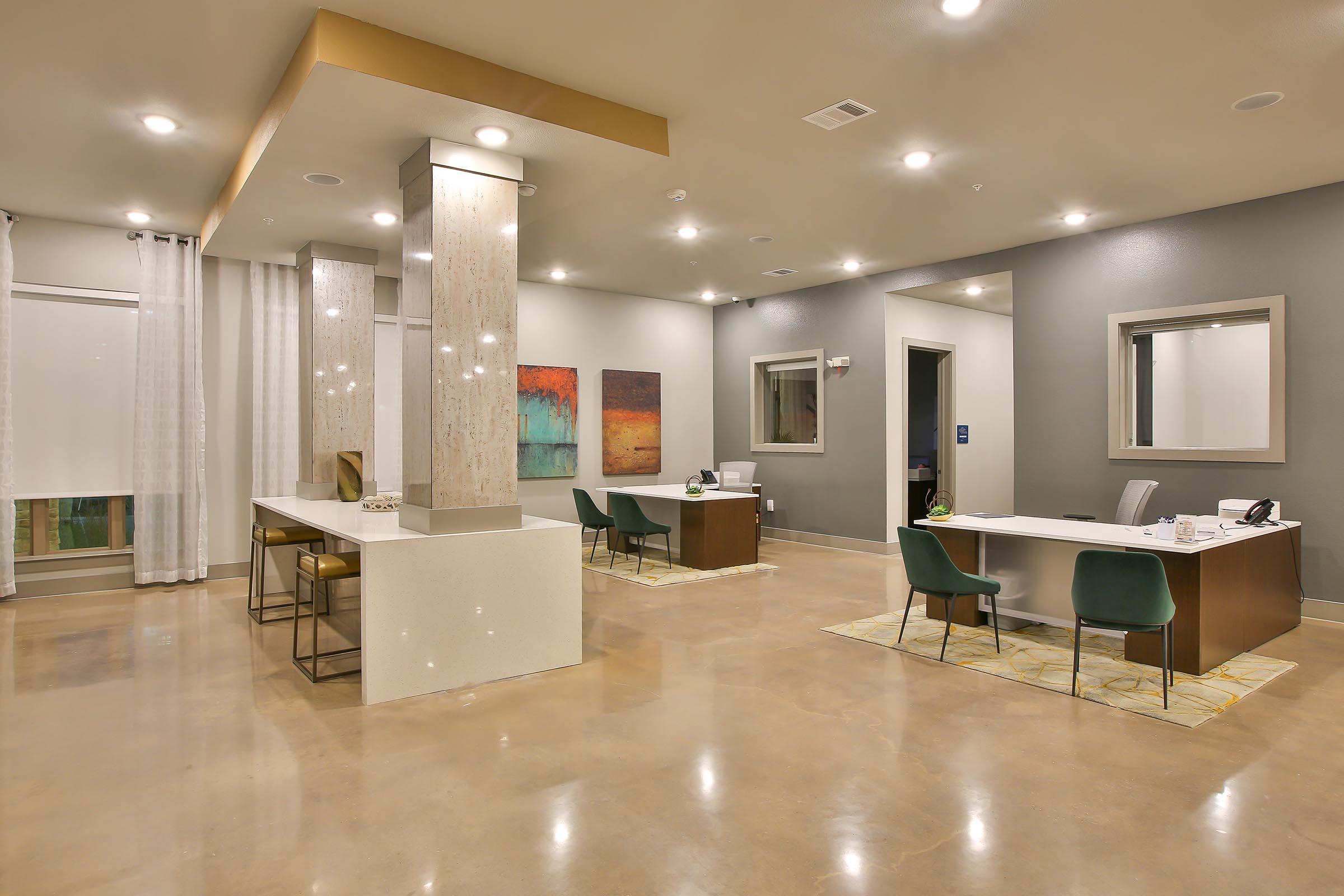 Modern interior of a reception area featuring sleek furnishings. Two desks are positioned around the space, with a neutral color palette and polished concrete floors. Abstract artwork adorns the walls, and large windows provide natural light, complemented by soft LED lighting overhead.