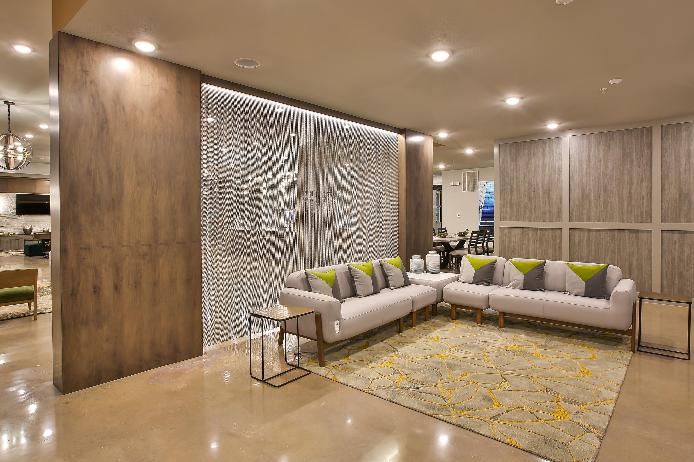 Modern lounge area featuring a light-colored sofa with gray and green cushions, a decorative rug with a geometric pattern, and a sleek side table. The backdrop includes a textured wall and an illuminated open space, creating a welcoming atmosphere.