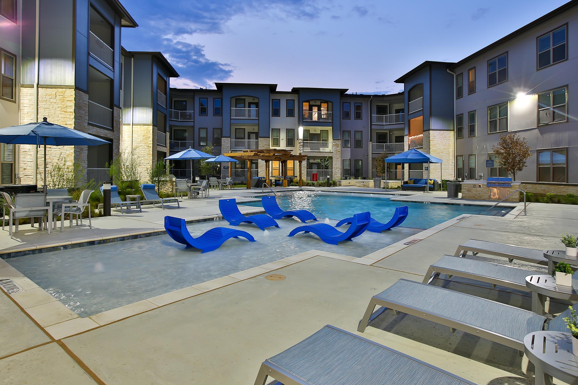 A modern apartment complex courtyard featuring a swimming pool with blue lounge chairs, surrounded by umbrellas and patio furniture. The building has multiple levels with balconies, and the scene is set during twilight with a clear sky.