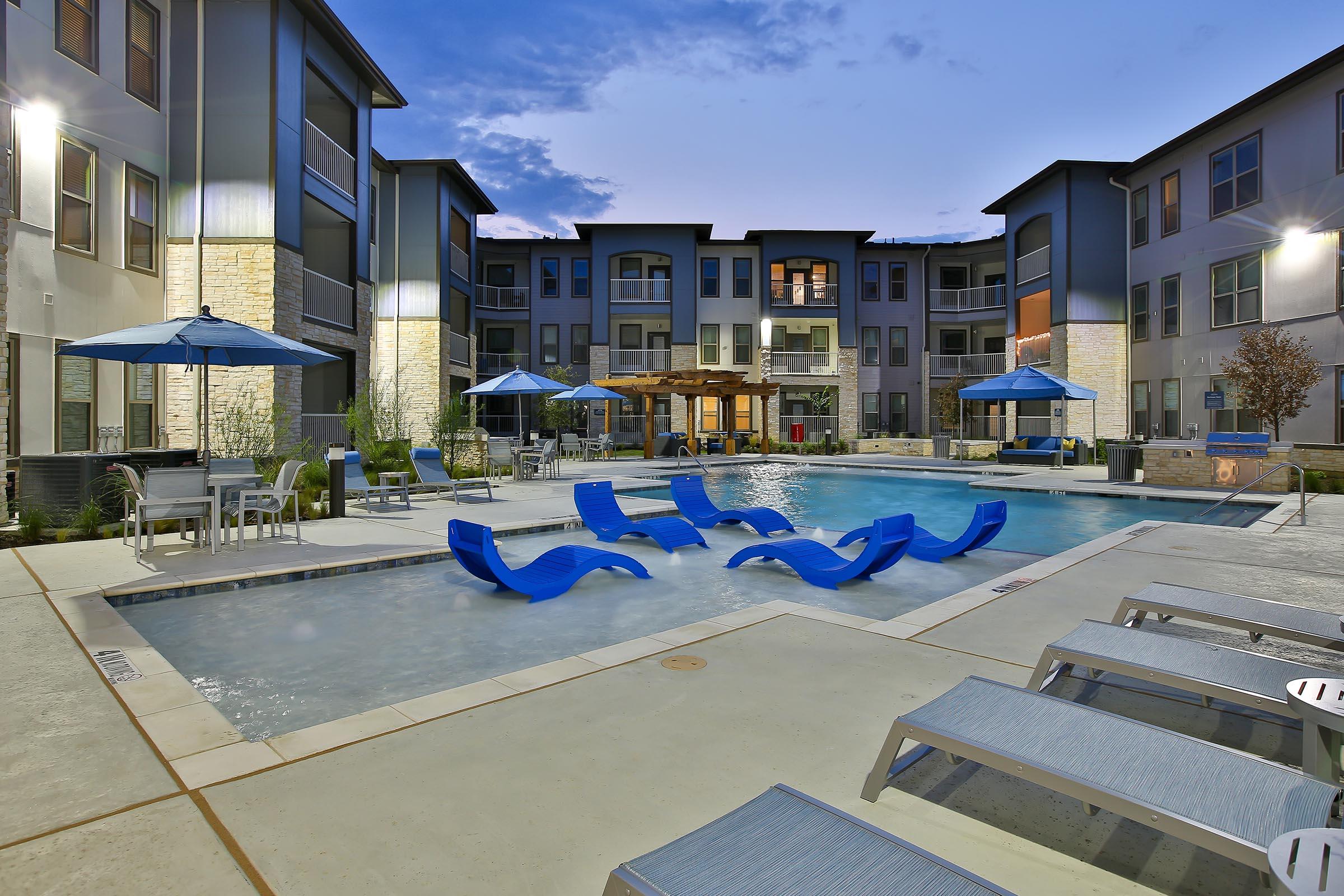 Modern apartment complex pool area at dusk, featuring a clear swimming pool surrounded by lounge chairs, blue umbrellas, and stylish architecture. The buildings have a contemporary design with large windows, and the scene is illuminated by soft lighting, creating a relaxing atmosphere.