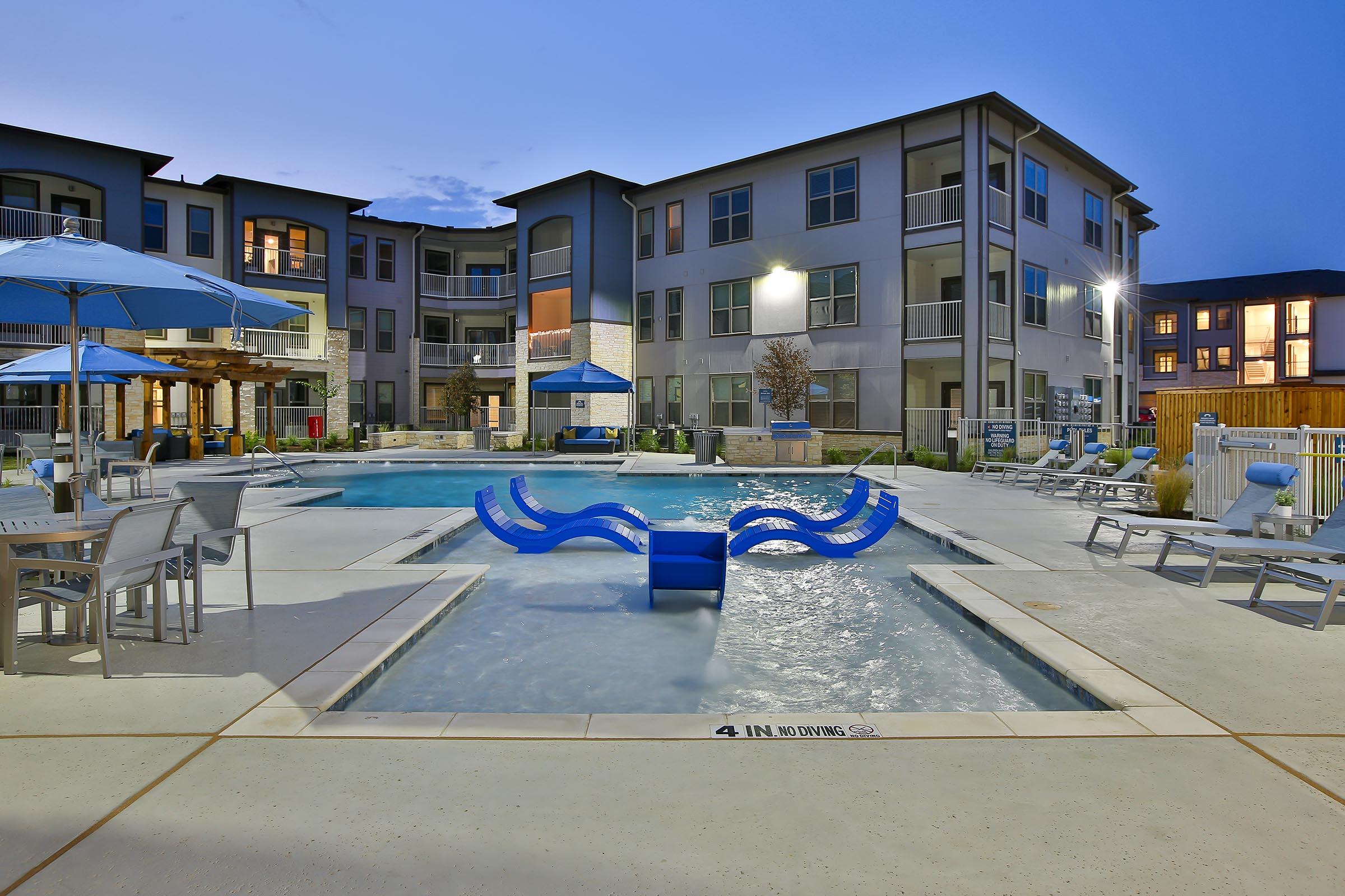 A modern apartment complex pool area at dusk. There are two swimming pools, one with a water feature, surrounded by lounge chairs and umbrellas. The buildings nearby have multiple stories and balconies, creating a welcoming outdoor space for residents.