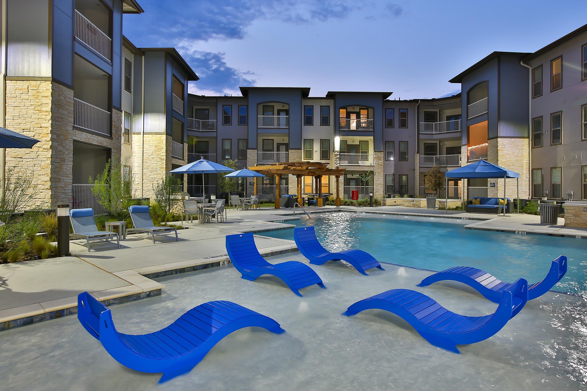 A modern apartment complex with a large swimming pool surrounded by blue lounge chairs and umbrellas. The buildings are stylish with a mix of stone and modern materials, set against a twilight sky. The pool area is designed for relaxation, with greenery adding to the inviting atmosphere.
