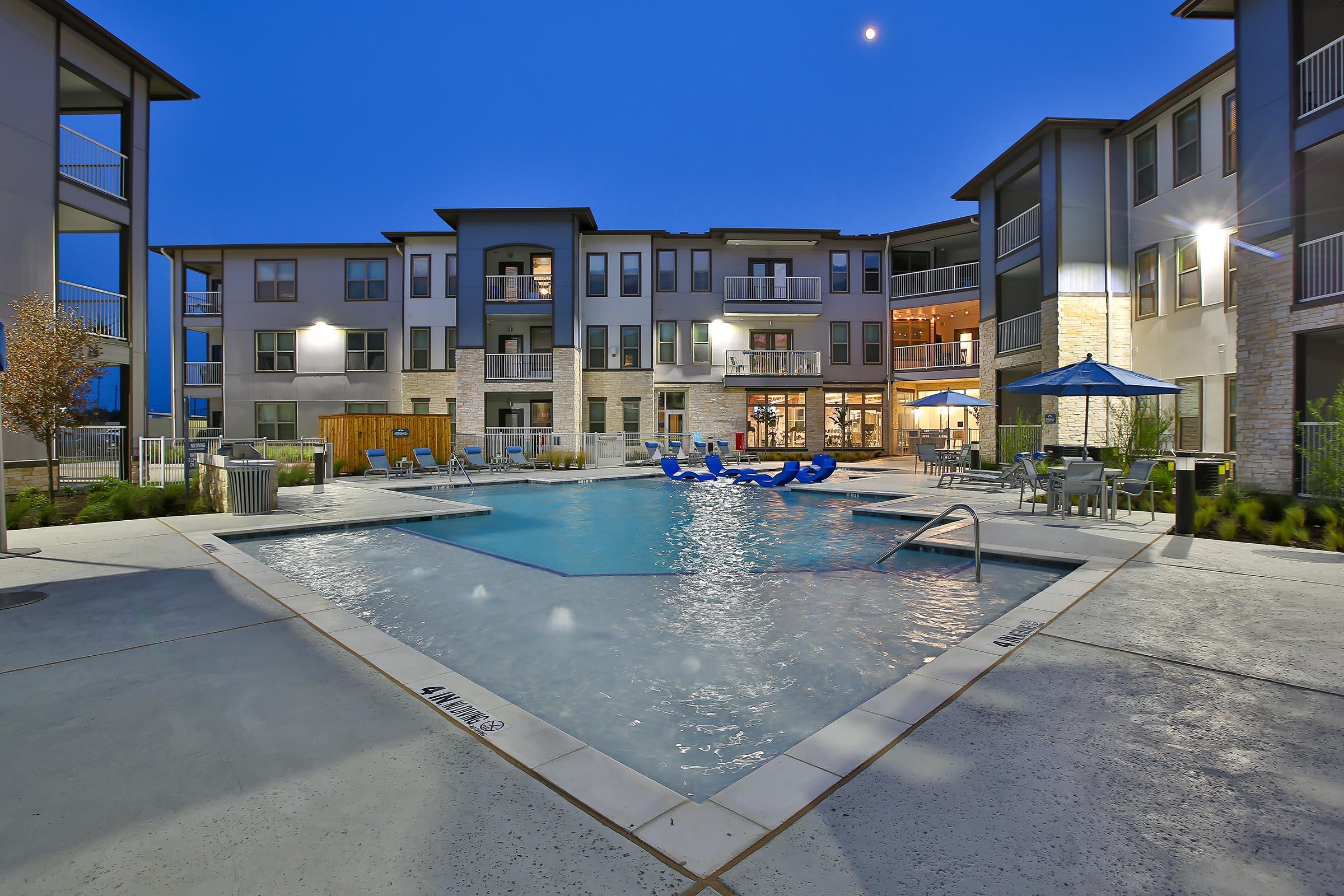 Modern apartment complex courtyard featuring a lit pool surrounded by lounge chairs, umbrellas, and well-maintained landscaping. The building, designed with contemporary architecture, has multiple balconies and large windows, creating a welcoming atmosphere in the evening light.