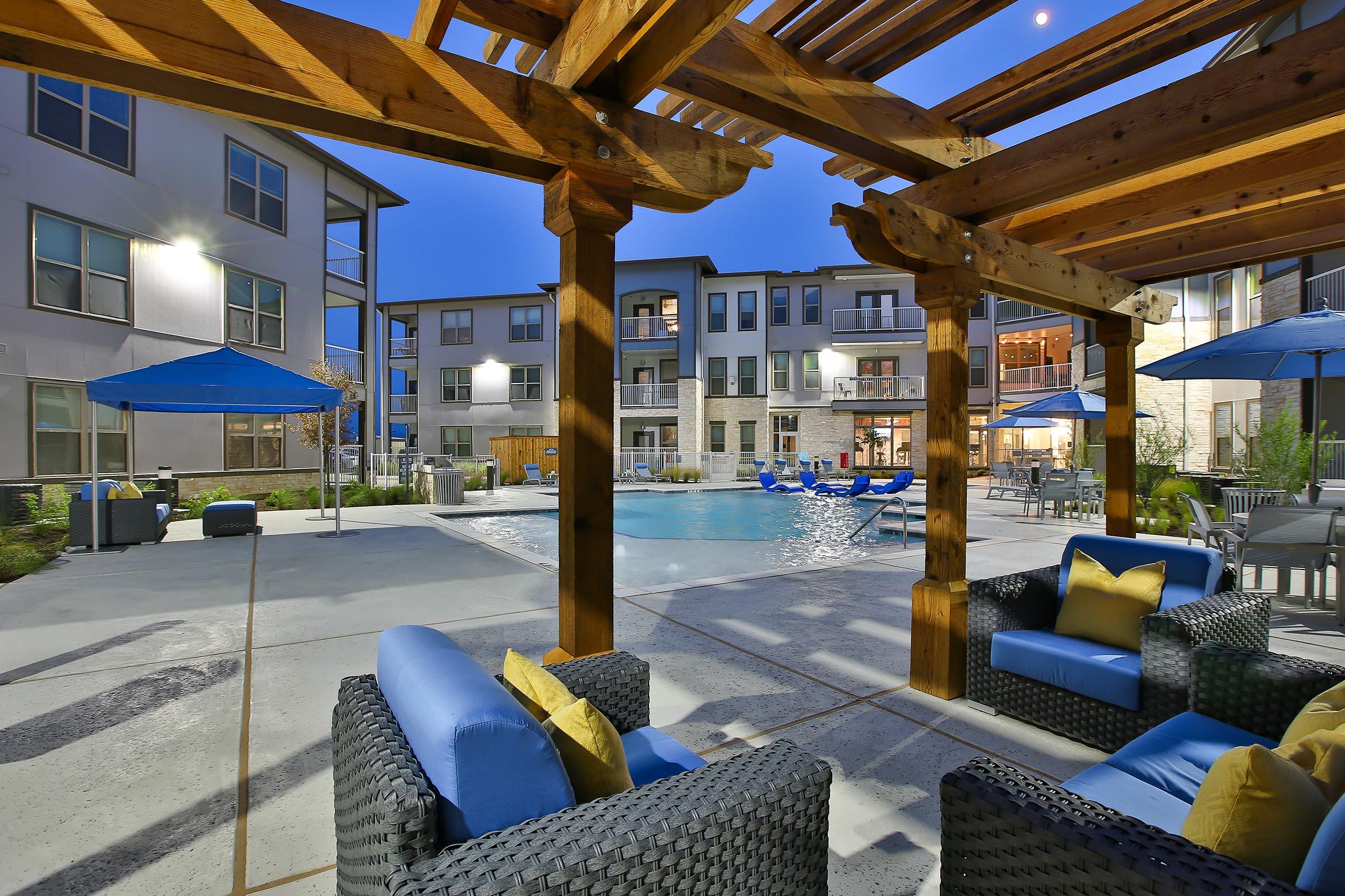 A cozy outdoor lounge area featuring comfortable seating with blue cushions and yellow throw pillows, overlooking a well-lit swimming pool. Surrounding the pool are umbrellas and patio furniture, with modern apartment buildings in the background, creating a relaxing evening atmosphere.