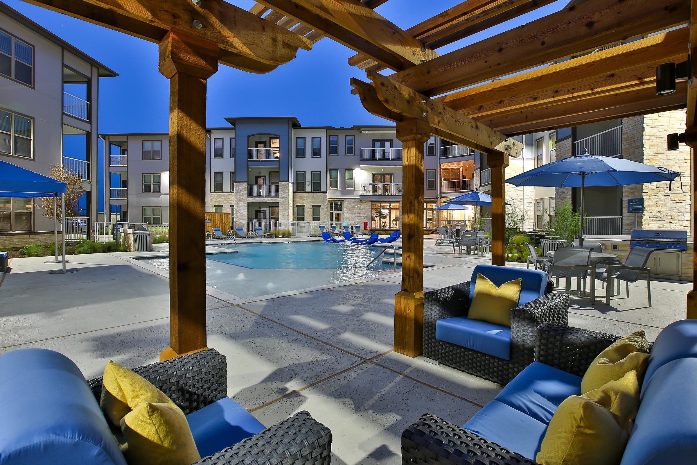 A stylish outdoor lounge area with blue cushioned chairs and yellow pillows, featuring a large swimming pool surrounded by buildings. There are umbrellas for shade and a warm evening ambiance, creating an inviting atmosphere for relaxation.