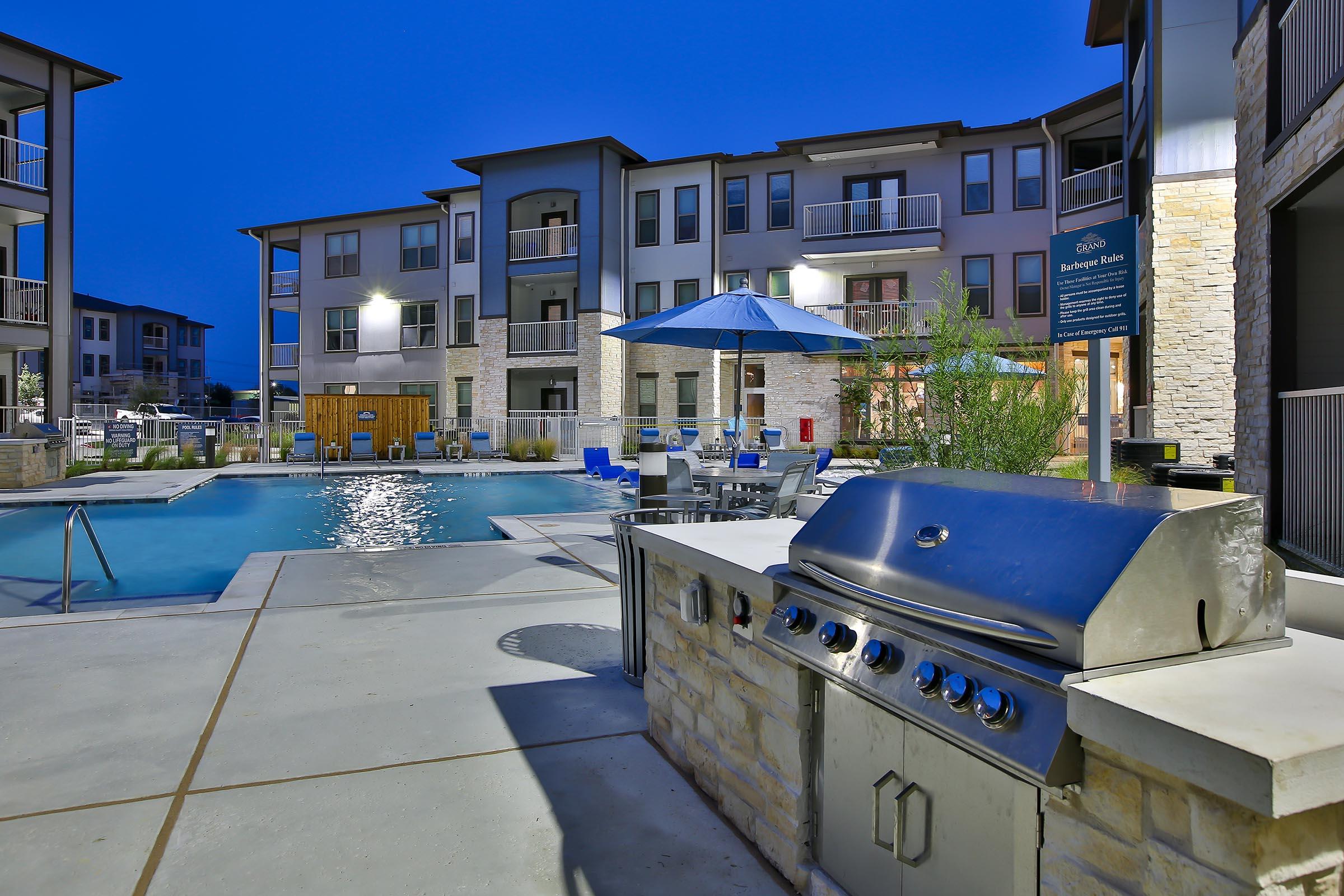 A modern outdoor space featuring a swimming pool surrounded by lounge chairs and tables. In the foreground, a stainless steel grill is set up near the pool area, with a blue umbrella providing shade. The scene is illuminated by soft lighting at dusk, creating a welcoming atmosphere for relaxation and socializing.