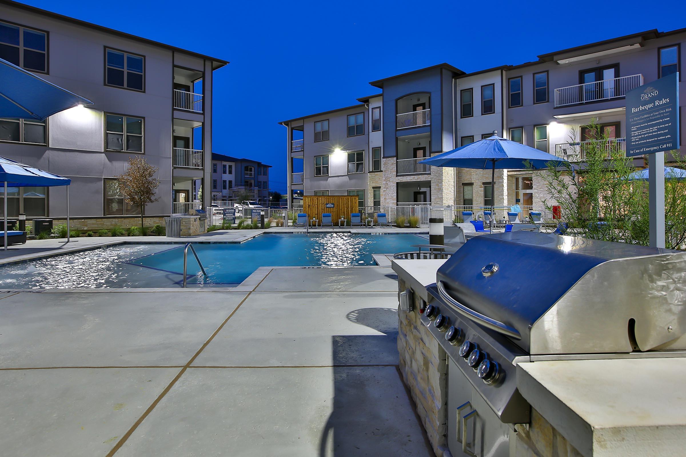 A beautifully designed outdoor pool area at dusk, featuring a modern swimming pool surrounded by lounge chairs and umbrellas. In the foreground, a stainless steel barbecue grill is positioned near the pool, with well-lit apartment buildings in the background.