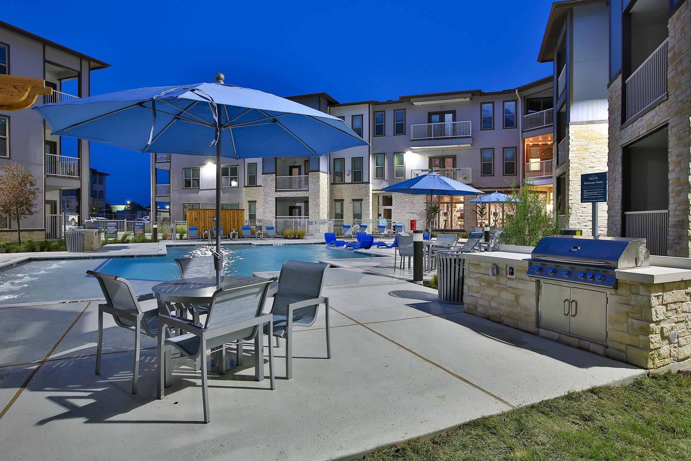 A well-lit apartment complex pool area at dusk, featuring a swimming pool, blue umbrellas over tables, lounge chairs, and a barbecue grill. The surrounding buildings showcase modern architecture, with balconies and windows reflecting the evening sky.