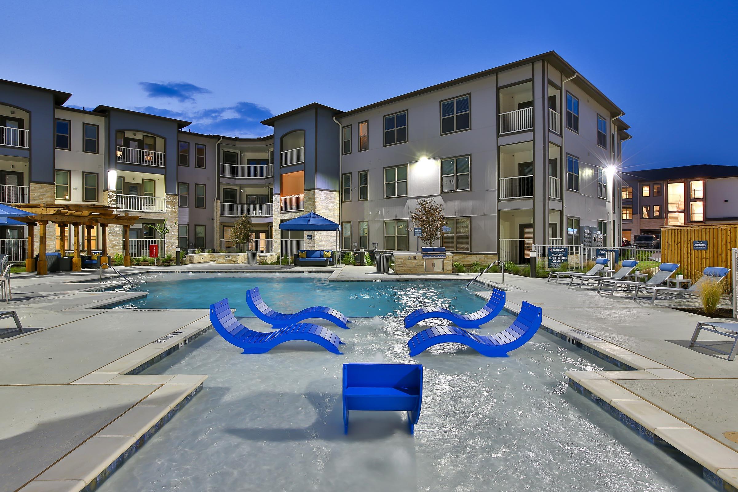 A modern apartment complex featuring a resort-style pool area at dusk. The pool has blue lounge chairs partially submerged in the water, surrounded by concrete decking and landscaped areas. Soft lighting enhances the inviting ambiance, with buildings in the background.
