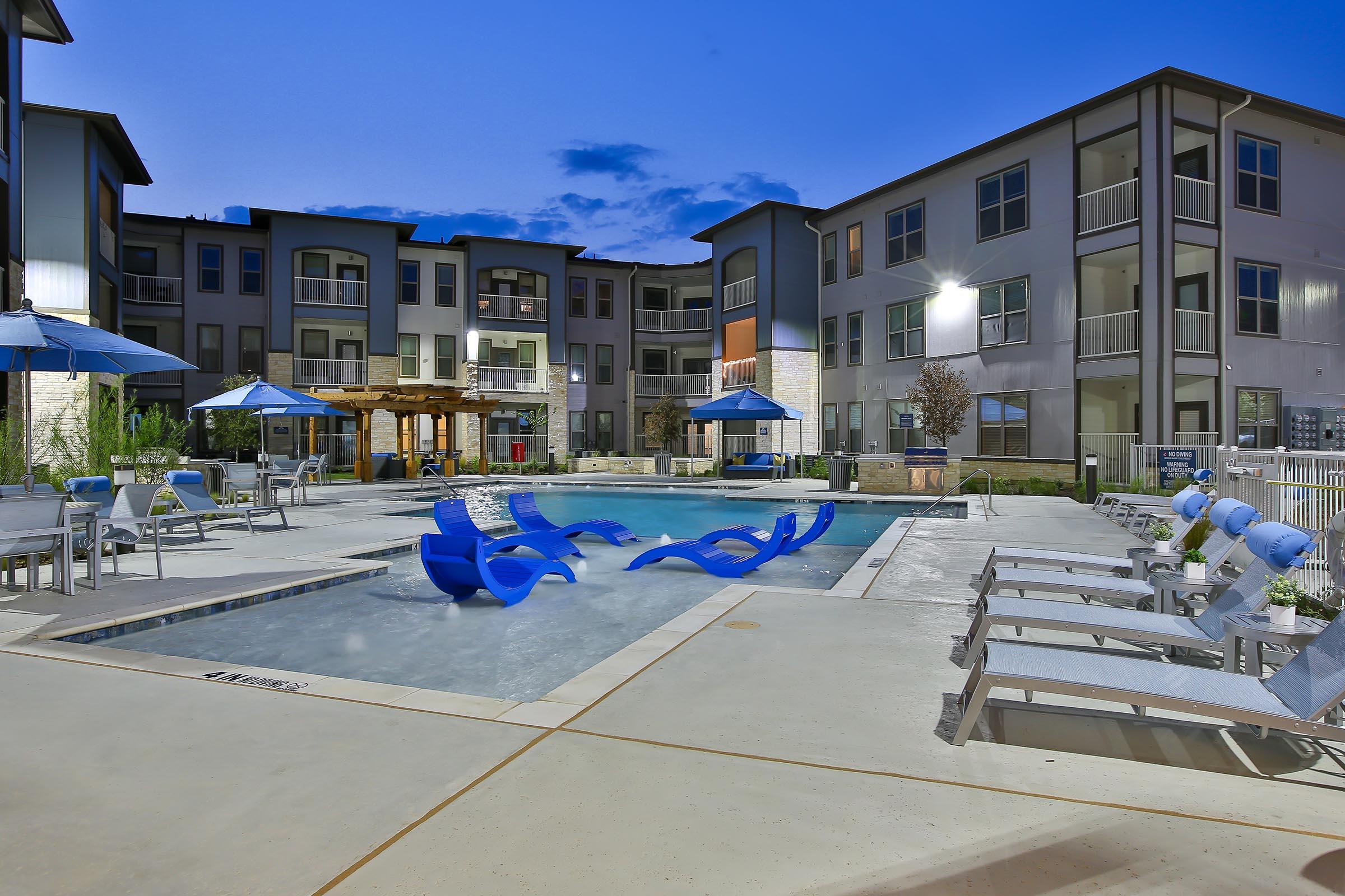 A modern outdoor pool area at an apartment complex, featuring blue lounge chairs, umbrellas, and a wooden pergola. The pool reflects the evening sky, and surrounding buildings showcase contemporary architecture. Soft lighting illuminates the area, creating a welcoming atmosphere for relaxation.