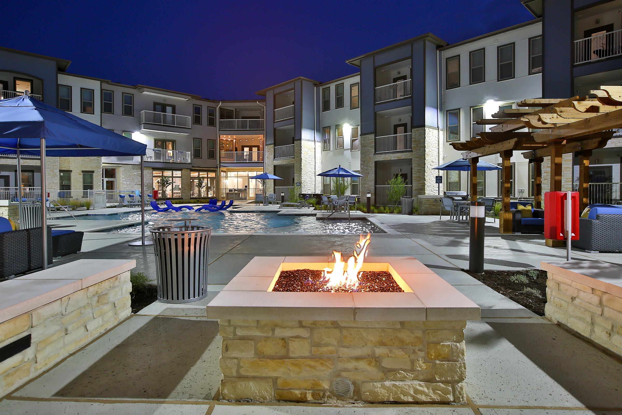 A modern apartment complex pool area at night, featuring a lit fire pit surrounded by stone seating, blue umbrellas, and lounge chairs near a sparkling water pool. The ambiance is inviting with well-lit buildings and landscaped surroundings.