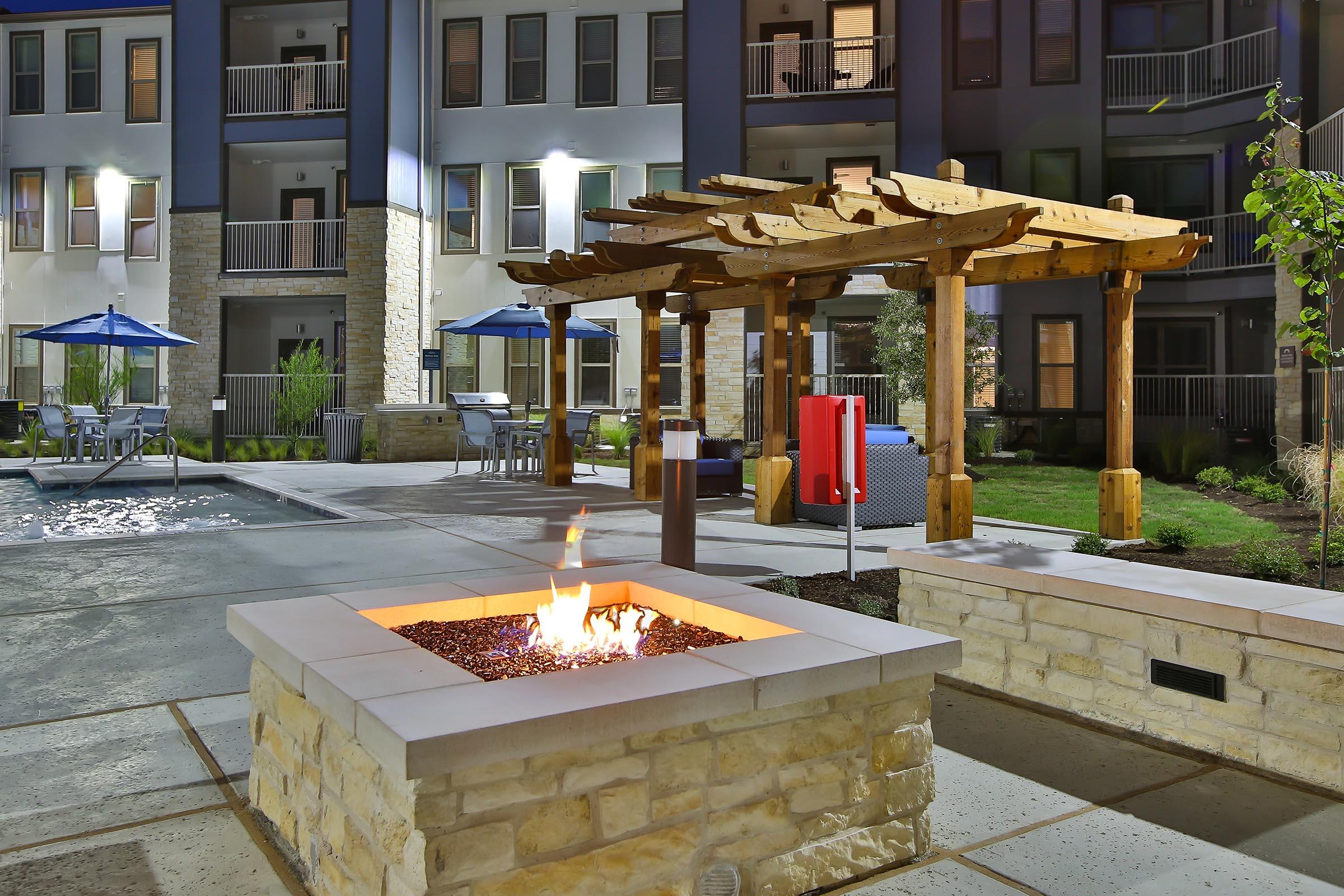 A well-lit outdoor area featuring a fire pit surrounded by pebbles, under a wooden pergola. In the background, there are lounge chairs and umbrellas near a pool, with modern apartment buildings and landscaping visible at night.