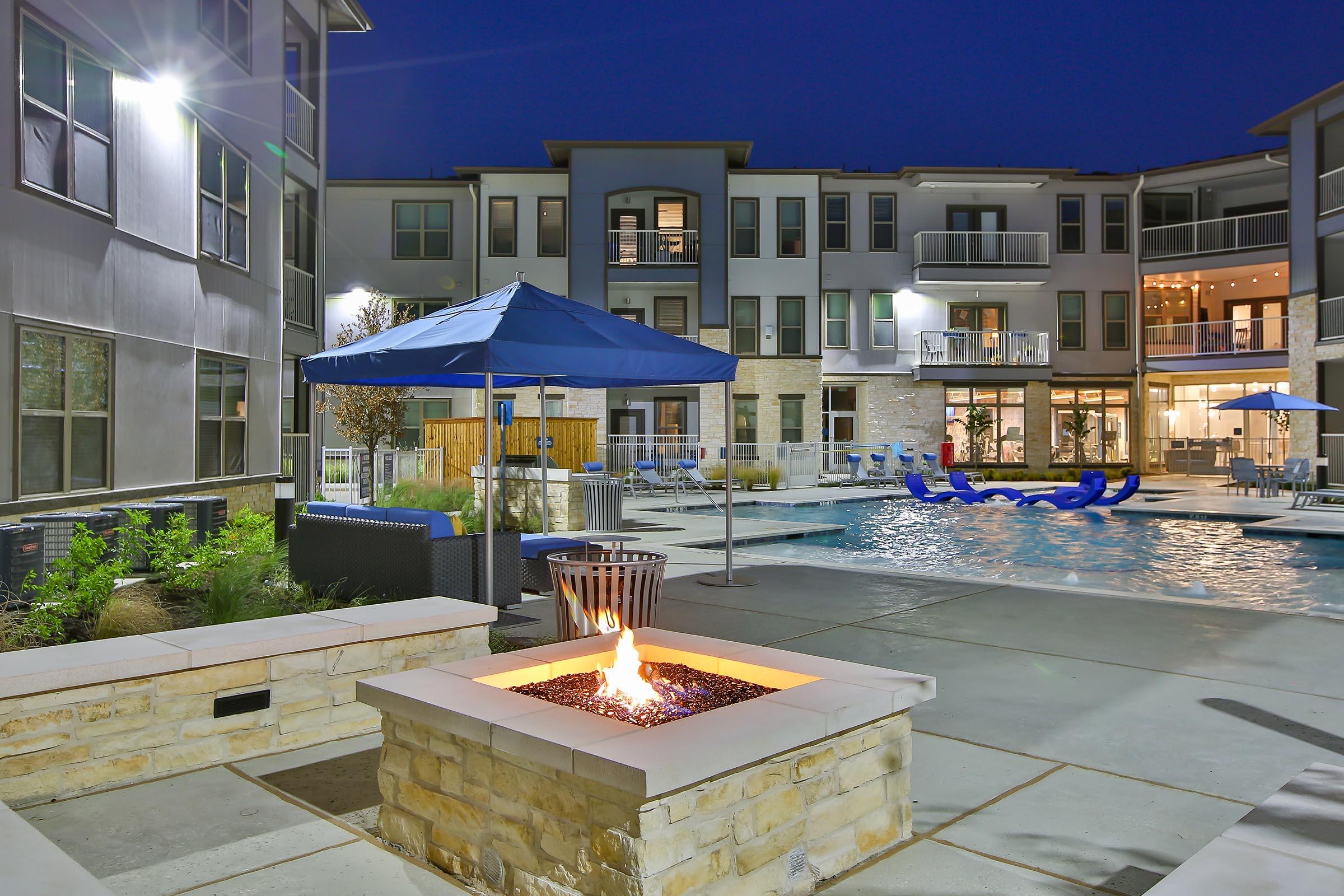 A modern apartment complex at night, featuring a courtyard with a swimming pool, blue lounge chairs, and a stone fire pit with a visible flame. There are several umbrellas around the pool area, and the buildings showcase large windows illuminated from within.