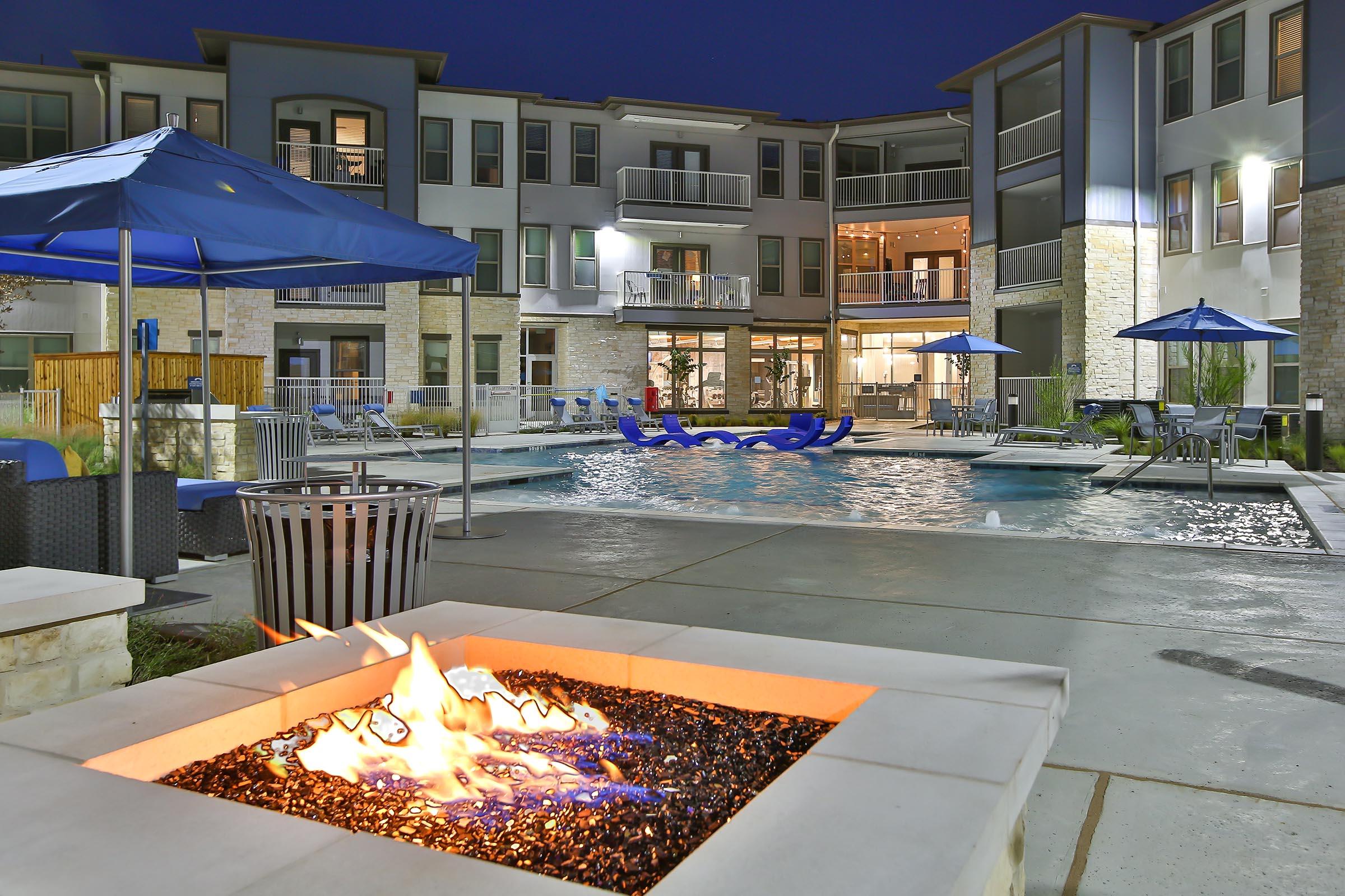 A modern outdoor pool area at dusk, featuring a fire pit with glowing embers in the foreground. Surrounding the pool are lounge chairs and umbrellas, with contemporary apartment buildings in the background. The scene is illuminated by soft lights, creating a relaxing atmosphere.