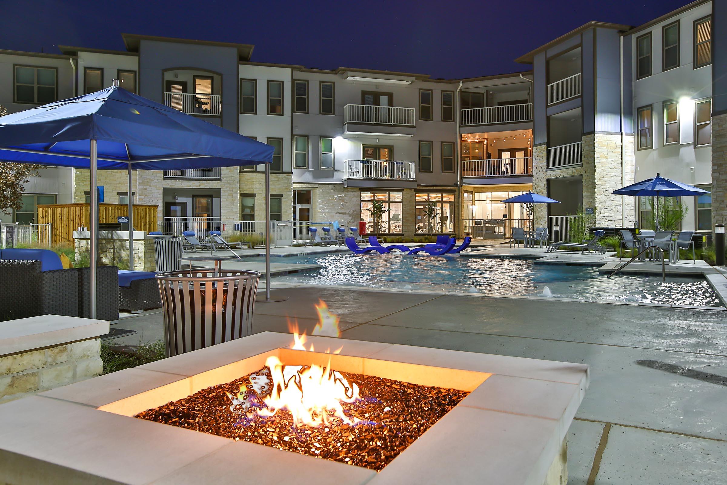 A modern apartment complex courtyard at night, featuring a swimming pool with lounge chairs, umbrellas, and a fire pit with flames. The building has multiple levels and large windows, reflecting a serene atmosphere. Lights from the pool and building create a warm ambiance.