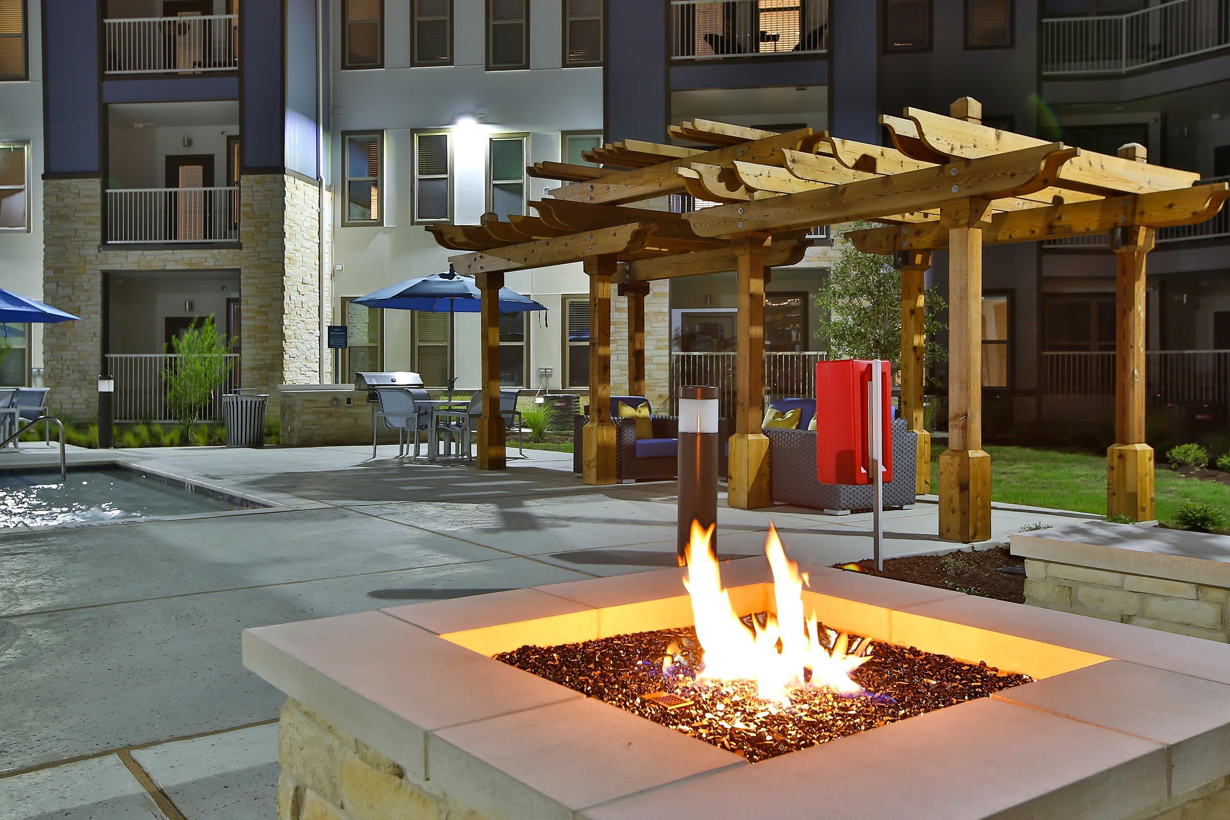 A cozy outdoor patio area at an apartment complex, featuring a fire pit with a flickering flame, surrounded by gravel. Nearby are wooden pergolas, colorful lounge furniture, and seating areas. In the background, the modern building is illuminated, creating a warm ambiance.