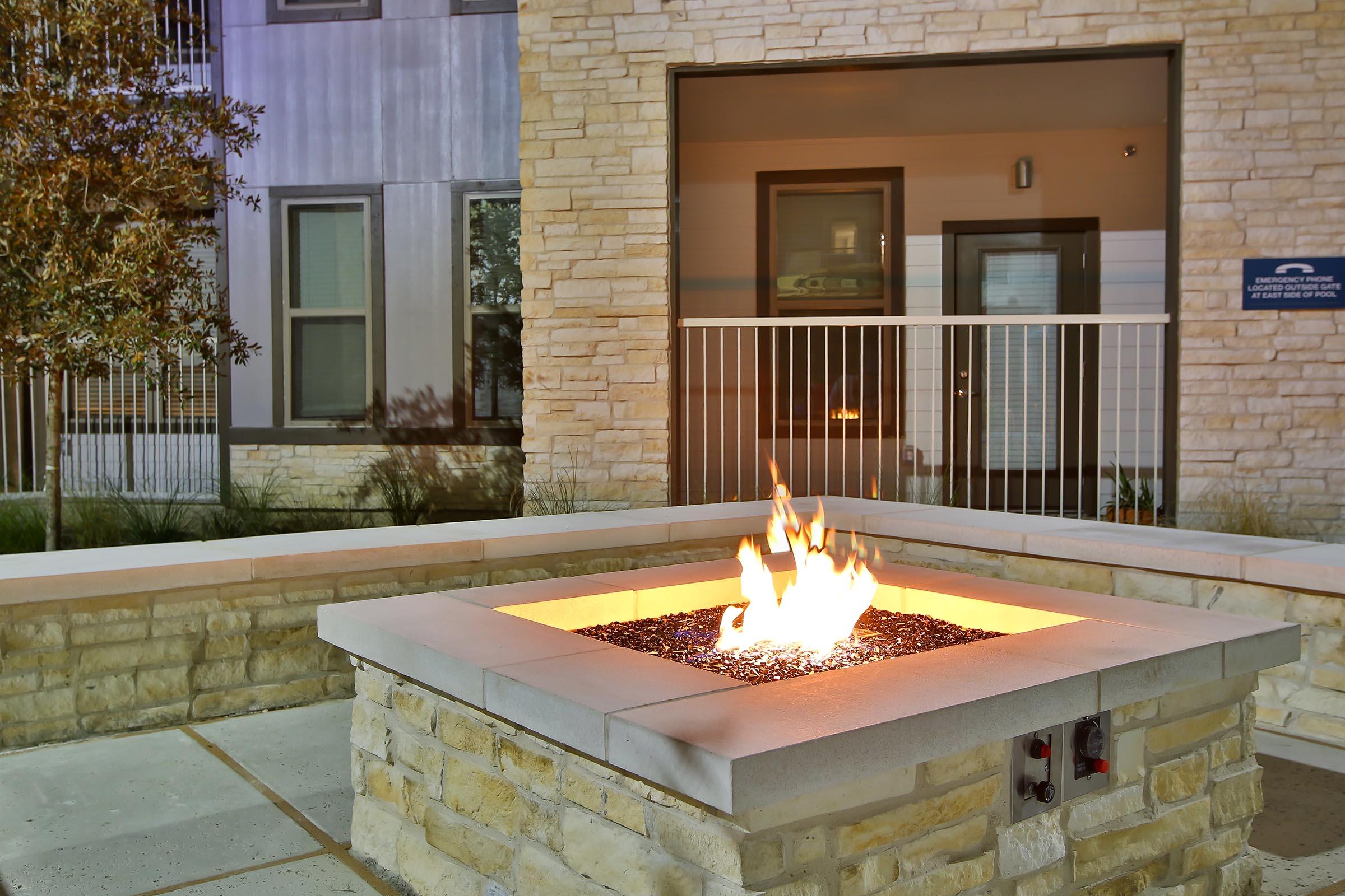 A modern outdoor fire pit made of stone, with a vibrant flame and black decorative glass stones. It is situated in a well-lit courtyard area, surrounded by contemporary architecture, with a green landscape in the background and a glimpse of an entrance to a building.