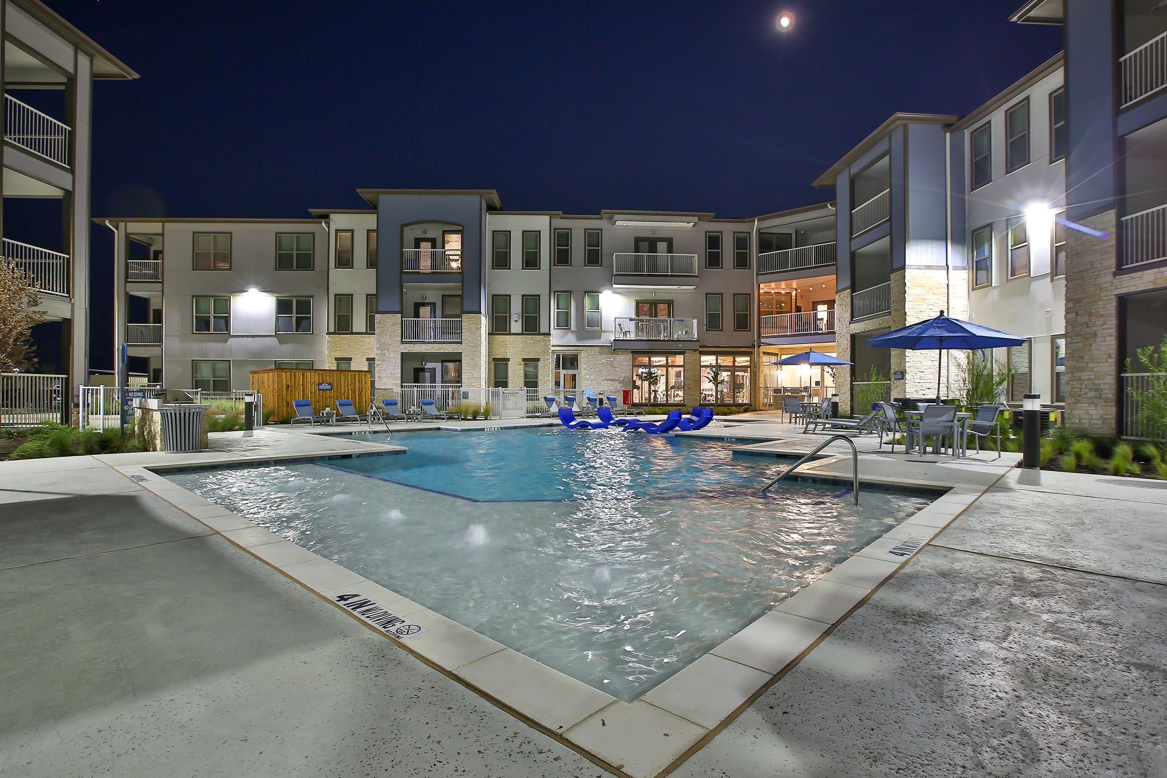 A nighttime view of a modern apartment complex featuring a well-lit swimming pool surrounded by lounge chairs. The building has multiple floors with balcony access, and there are umbrellas and seating areas nearby, all creating a welcoming atmosphere for residents and guests.