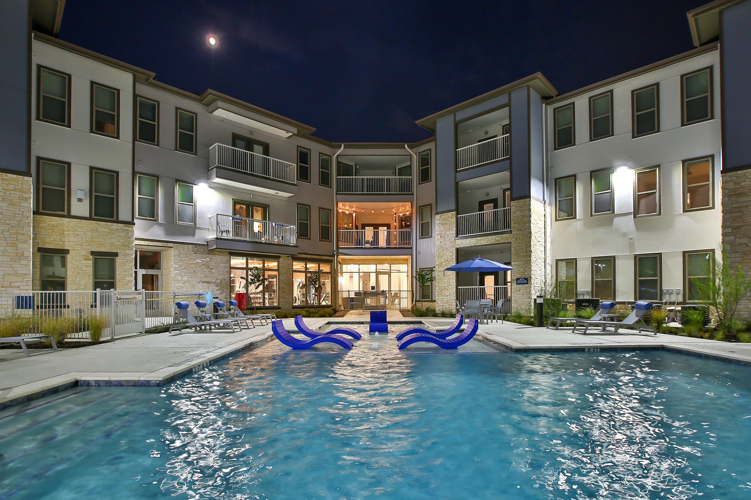 A well-lit apartment complex at night featuring a swimming pool in the foreground. The pool has lounge chairs around it, and the building showcases large windows and balconies. A moon is visible in the sky, adding to the tranquil ambiance of the scene.