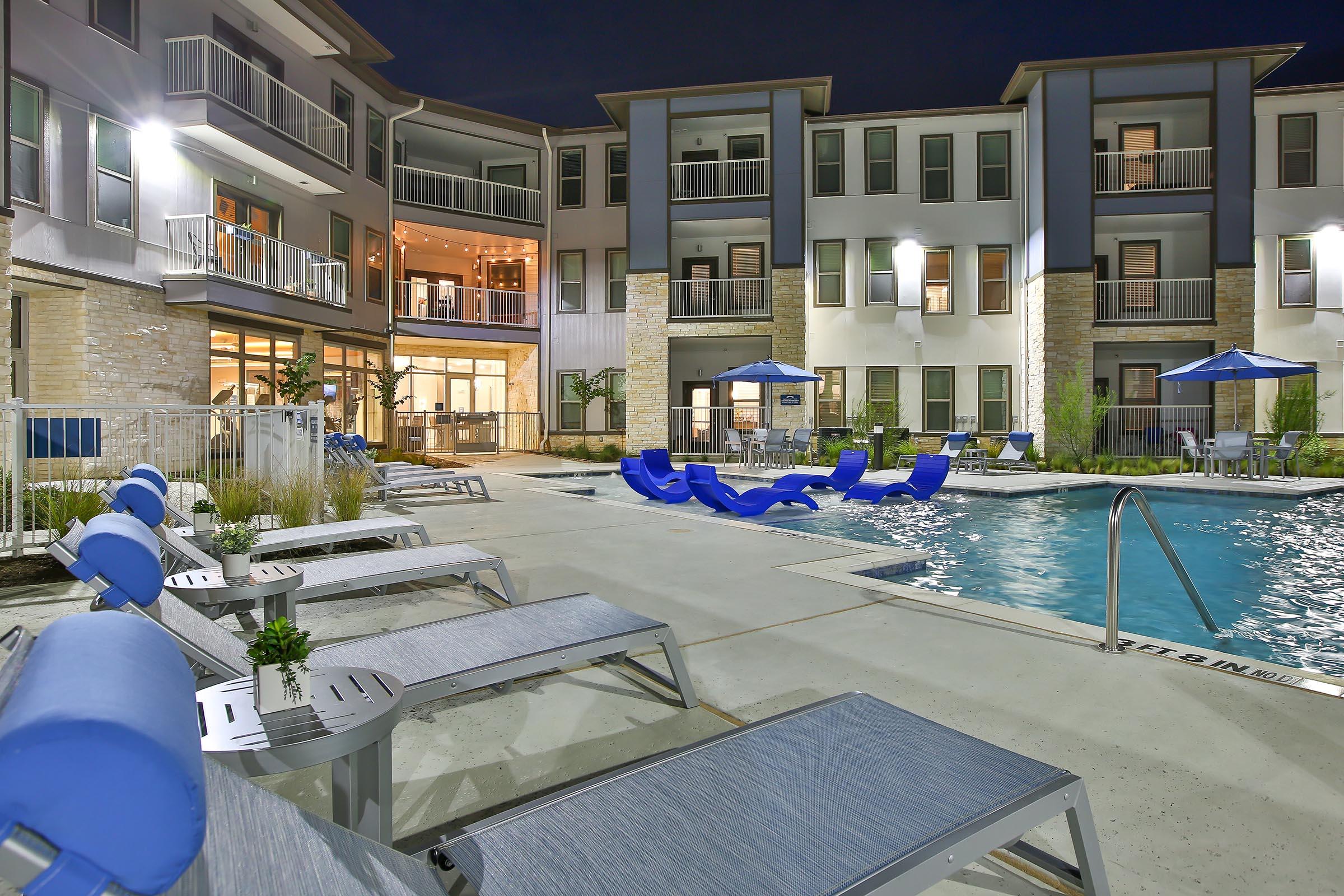 A modern apartment complex pool area at night, featuring lounge chairs and umbrellas beside a well-lit swimming pool. The building has multiple floors with balconies, and potted plants enhance the outdoor space. The atmosphere is inviting and stylish, ideal for relaxation and socializing.