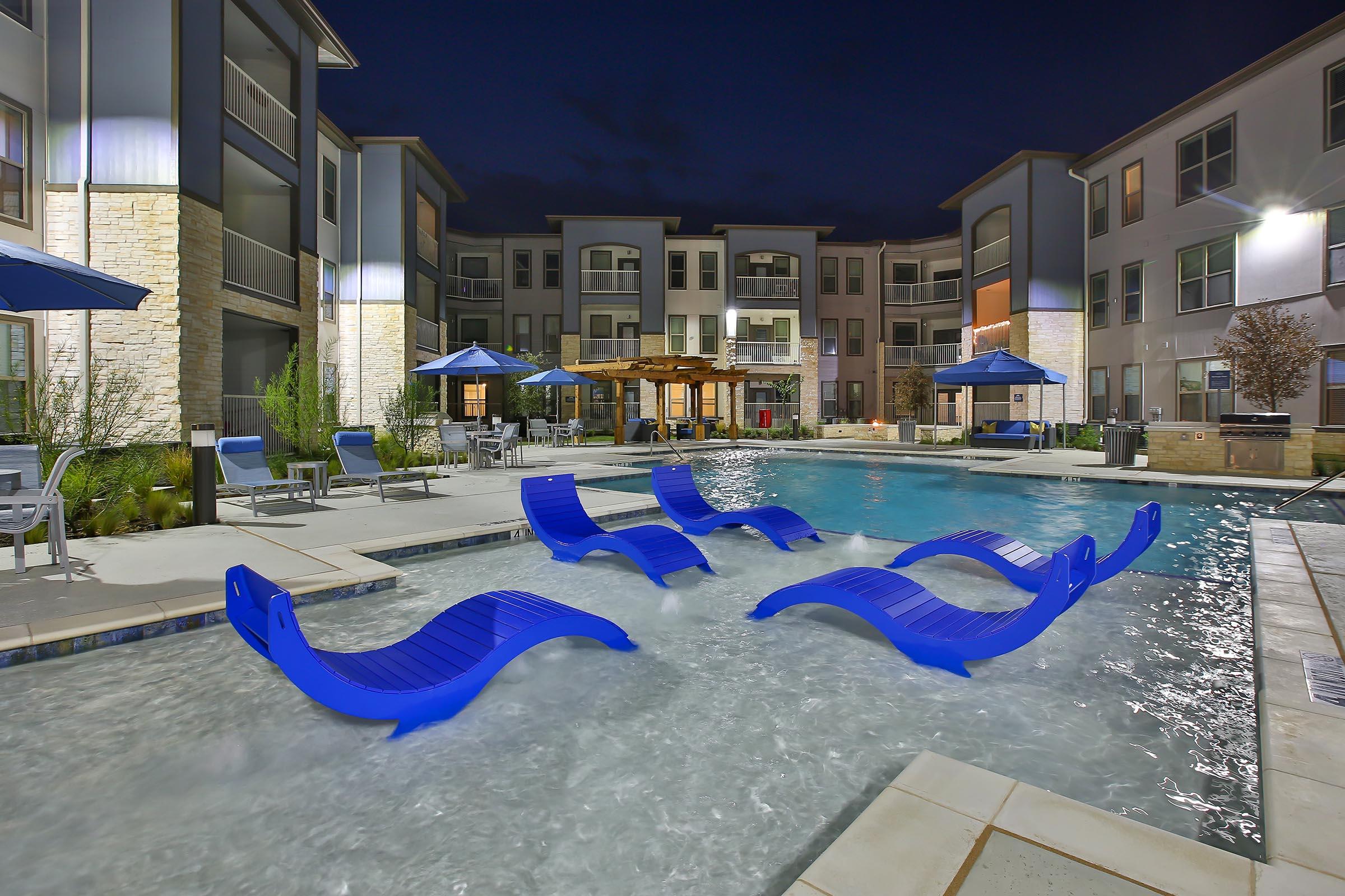 A nighttime view of a modern apartment complex featuring a swimming pool. The pool is surrounded by lounge chairs in blue, with shaded areas from umbrellas. The buildings are well-lit, highlighting their contemporary architecture. Lush landscaping adds to the serene atmosphere.