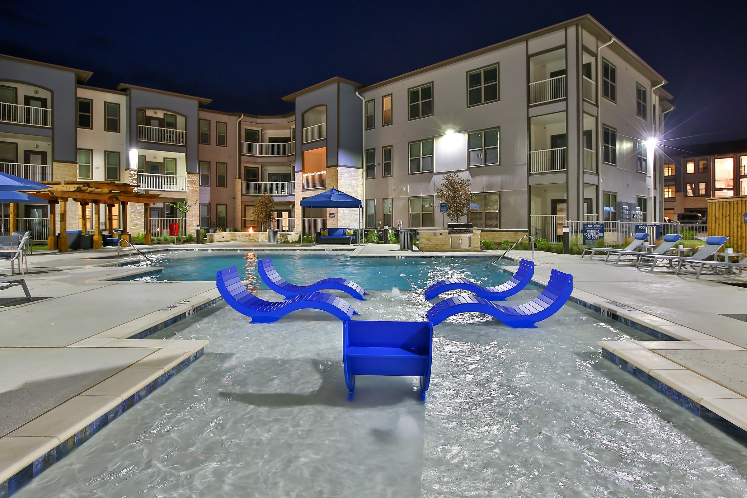 A vibrant apartment complex pool area at night, featuring a large swimming pool with illuminated water. In the center, there are blue lounge chairs partially submerged in the water, surrounded by comfortable seating and cabanas. The building has multiple balconies, adding to the modern aesthetic of the scene.