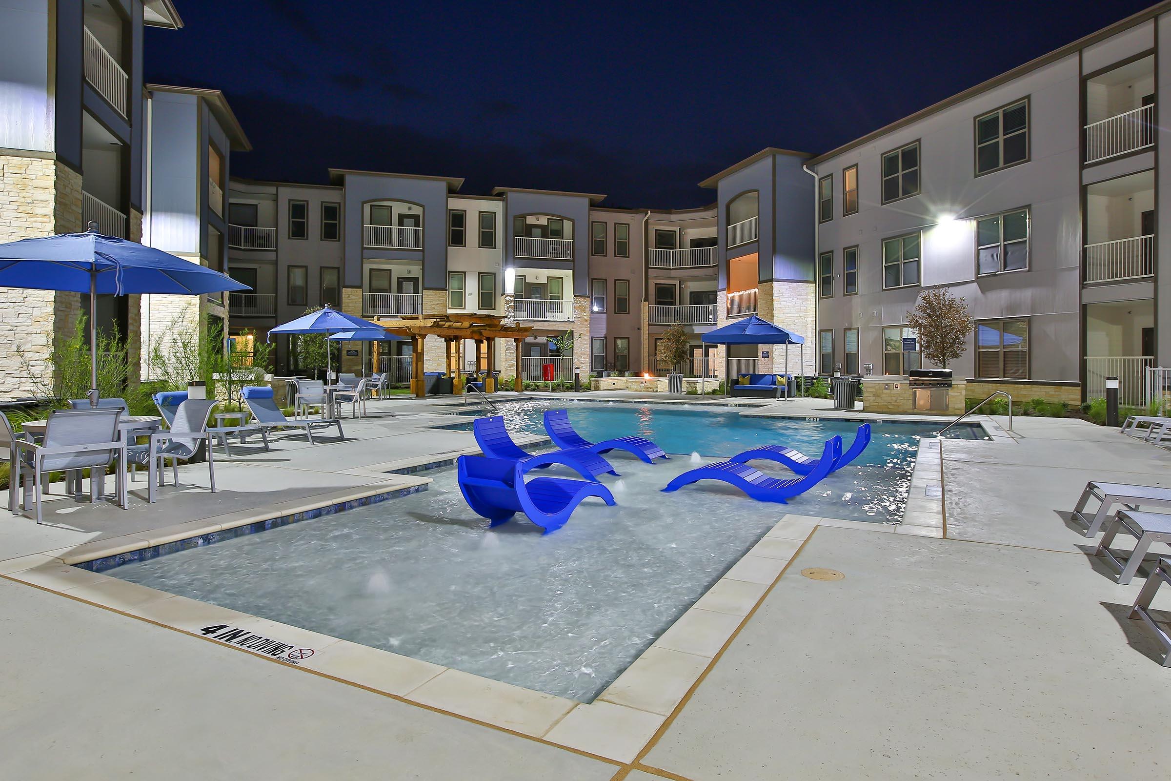 A nighttime view of a modern apartment complex pool area, featuring a swimming pool with blue lounge chairs partially submerged. Surrounding the pool are umbrellas and seating areas, with nearby buildings illuminated, creating a welcoming atmosphere.