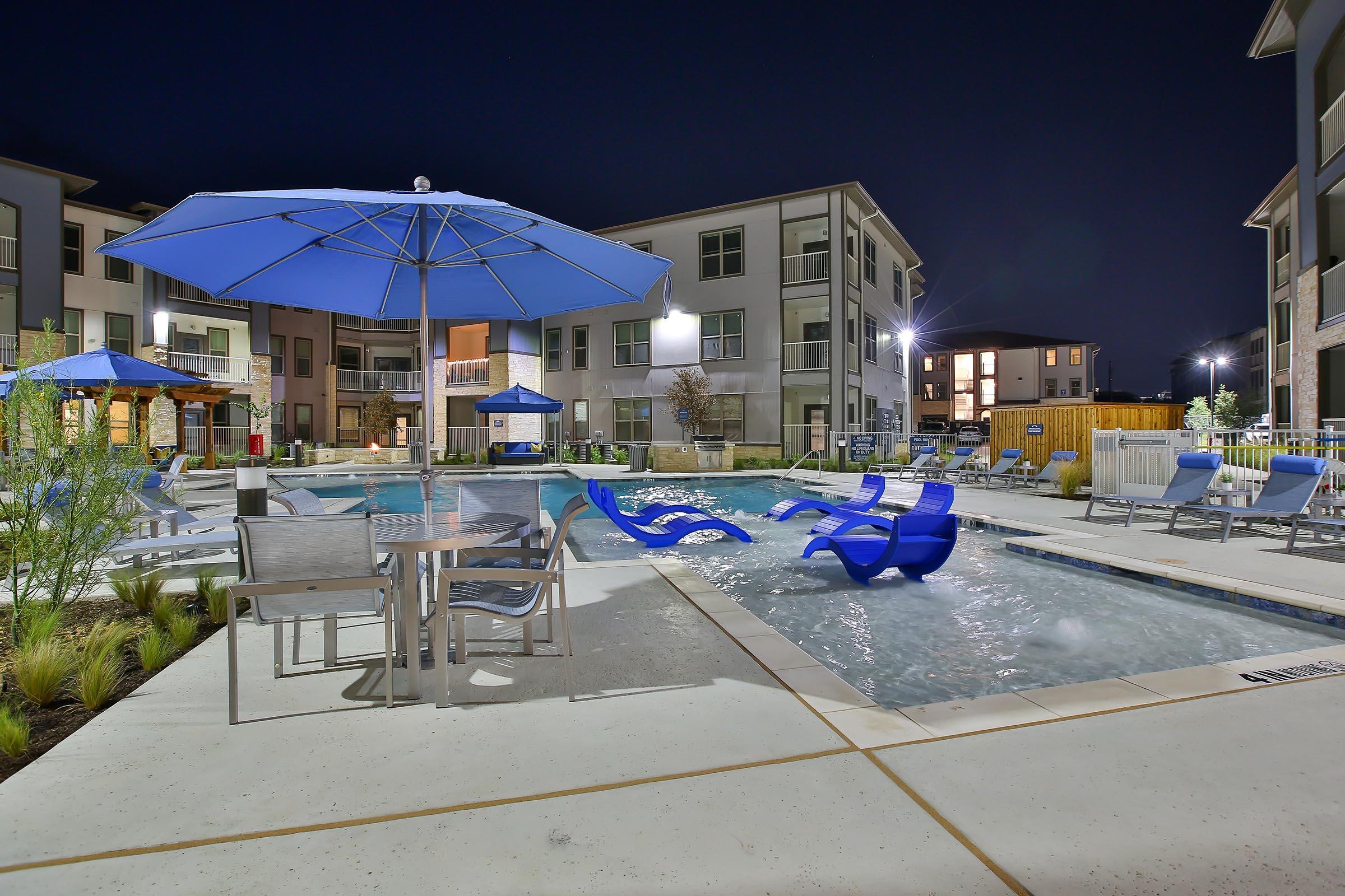 A nighttime view of a beautiful apartment complex pool area, featuring blue umbrellas, lounge chairs, and a bubbling spa. The surrounding landscape includes neatly trimmed grass and trees, with buildings lit in the background, creating a relaxing atmosphere.