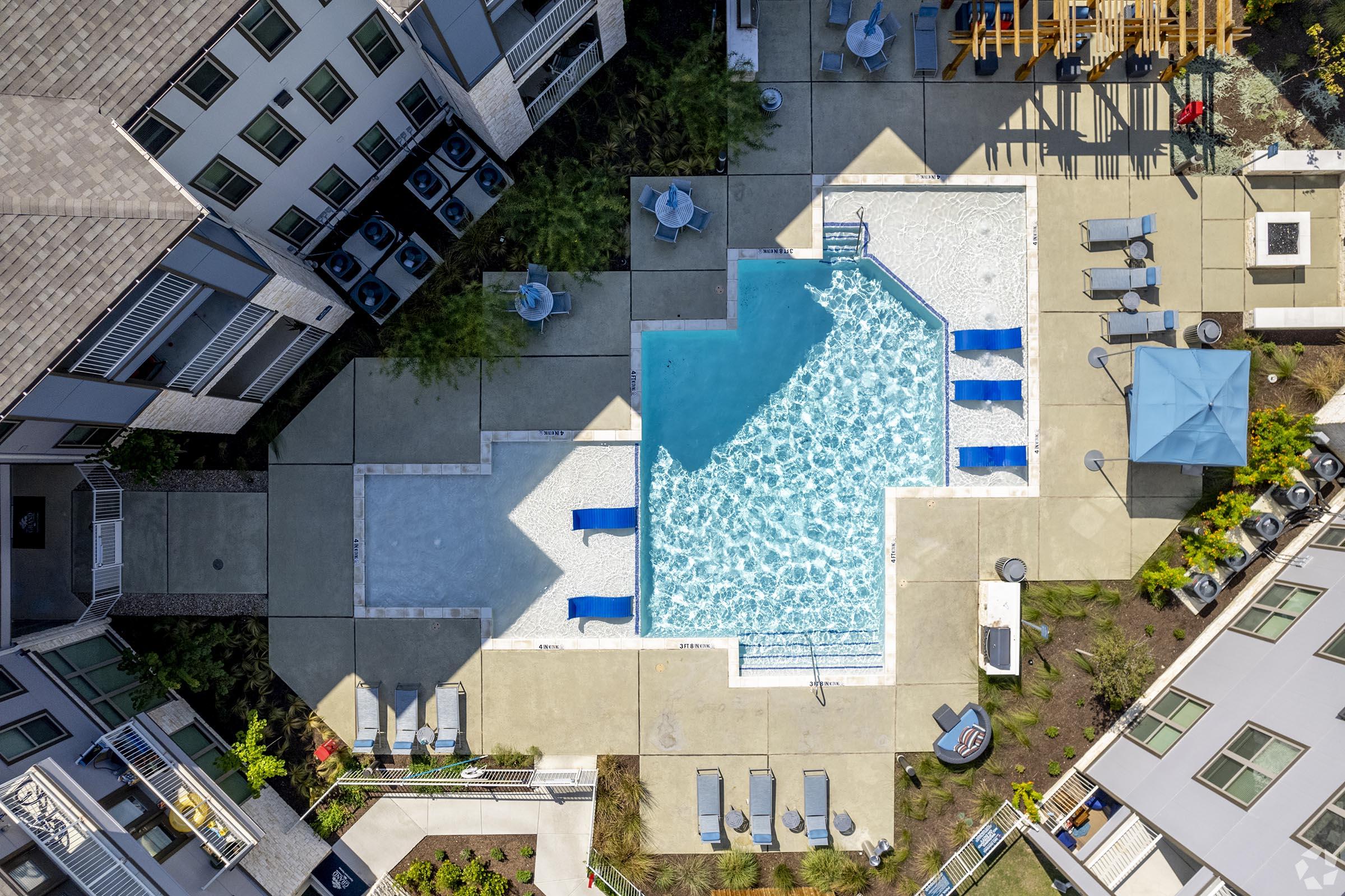 Aerial view of a residential pool area featuring a large swimming pool with blue lounge chairs around it. Nearby is a smaller pool, surrounded by a landscaped courtyard that includes seating areas and sunshades, with modern apartment buildings in the background.