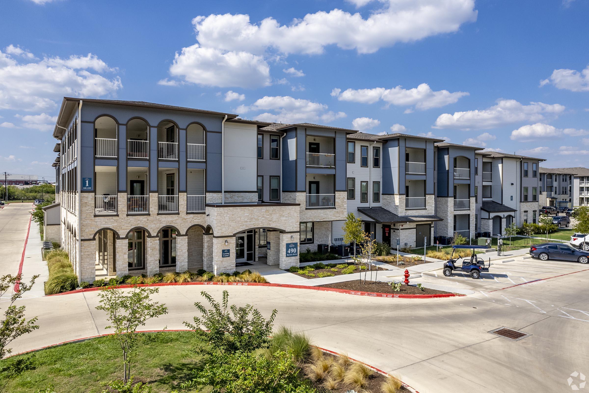 Modern multi-story apartment building with a mix of brick and stucco exteriors, featuring balconies and landscaped surroundings. The scene includes a well-maintained driveway and parking area, with blue skies and fluffy clouds in the background, creating a welcoming residential atmosphere.